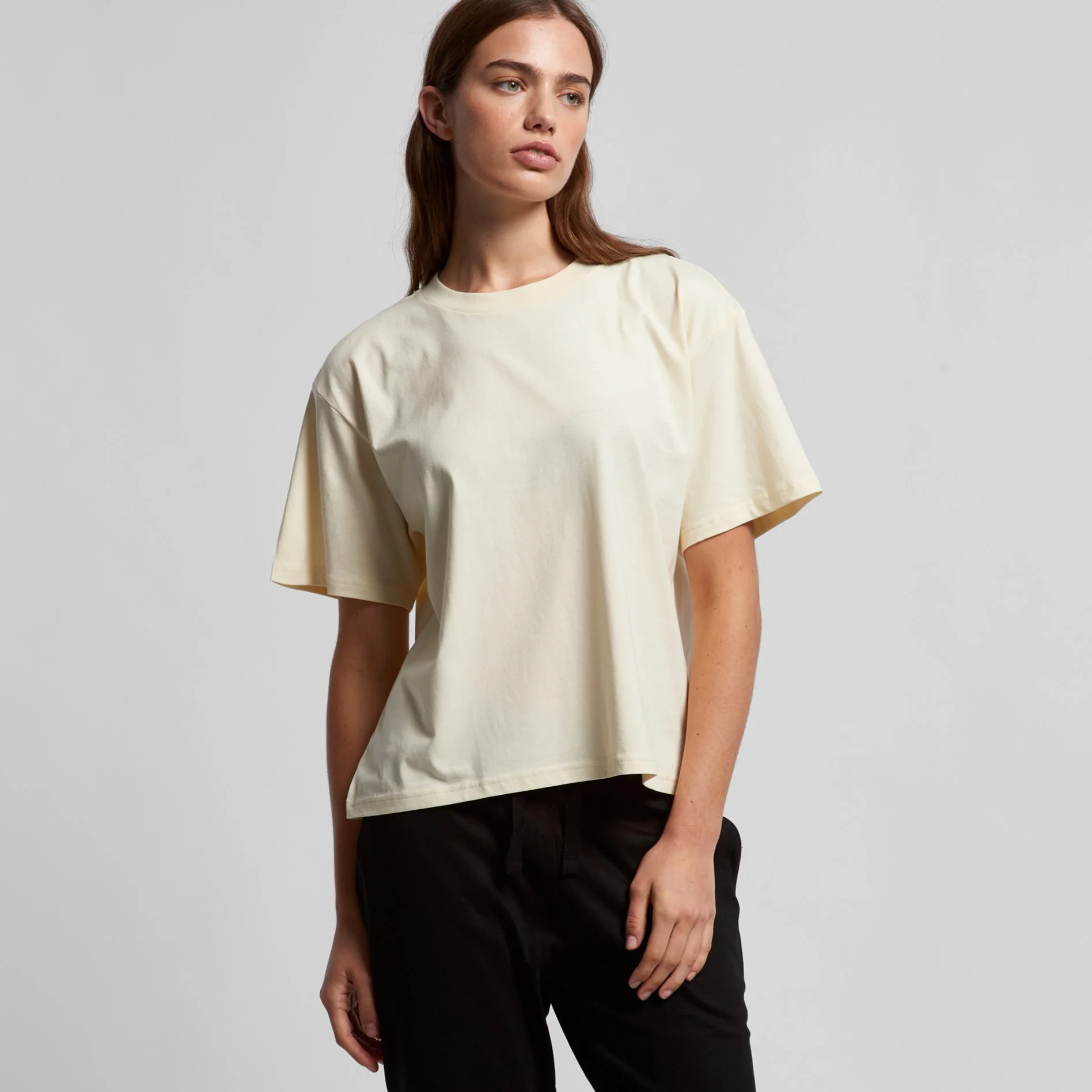 A woman with long brown hair and fair skin wearing a loose-fitting, cream-colored T-shirt and black pants, standing against a plain, light gray background.