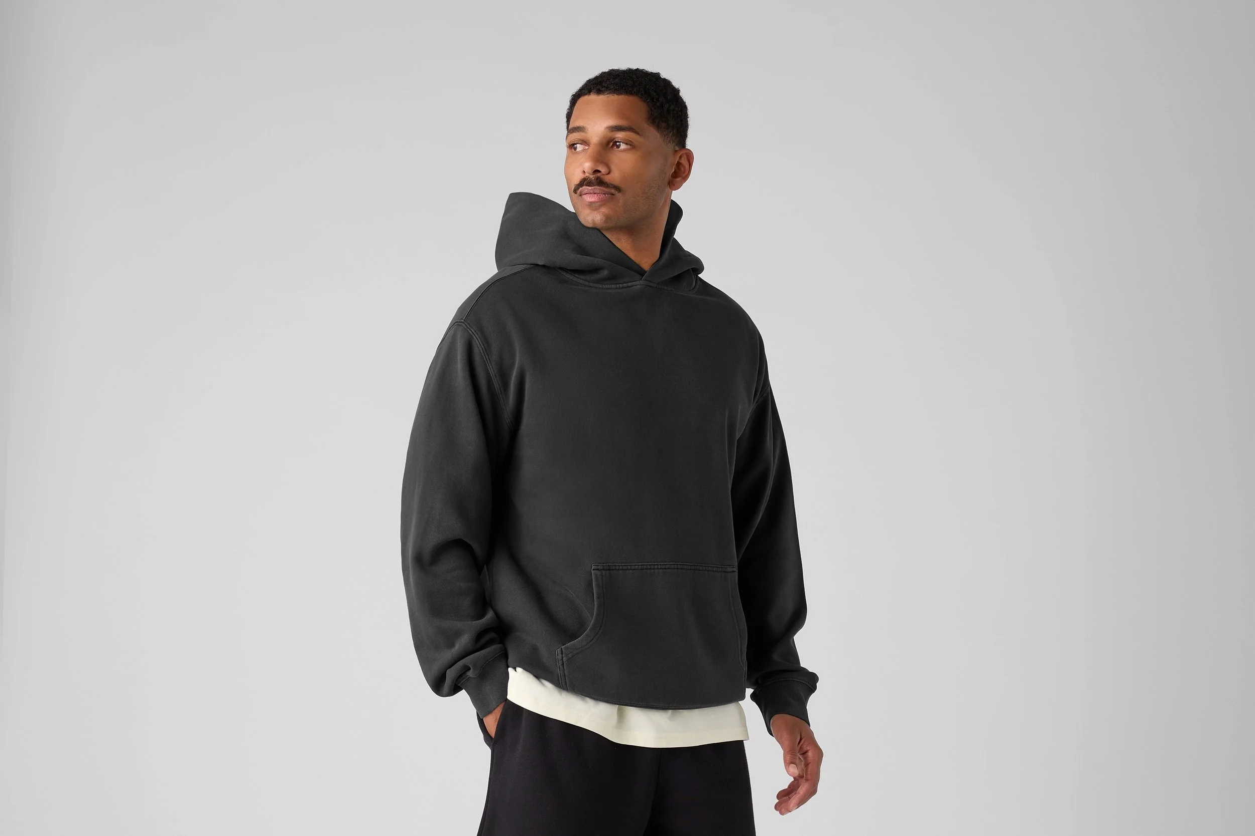 A young man wearing a black hoodie with a front pocket, standing against a light gray background. He has short, curly hair and a mustache, and his hands are in his pockets.