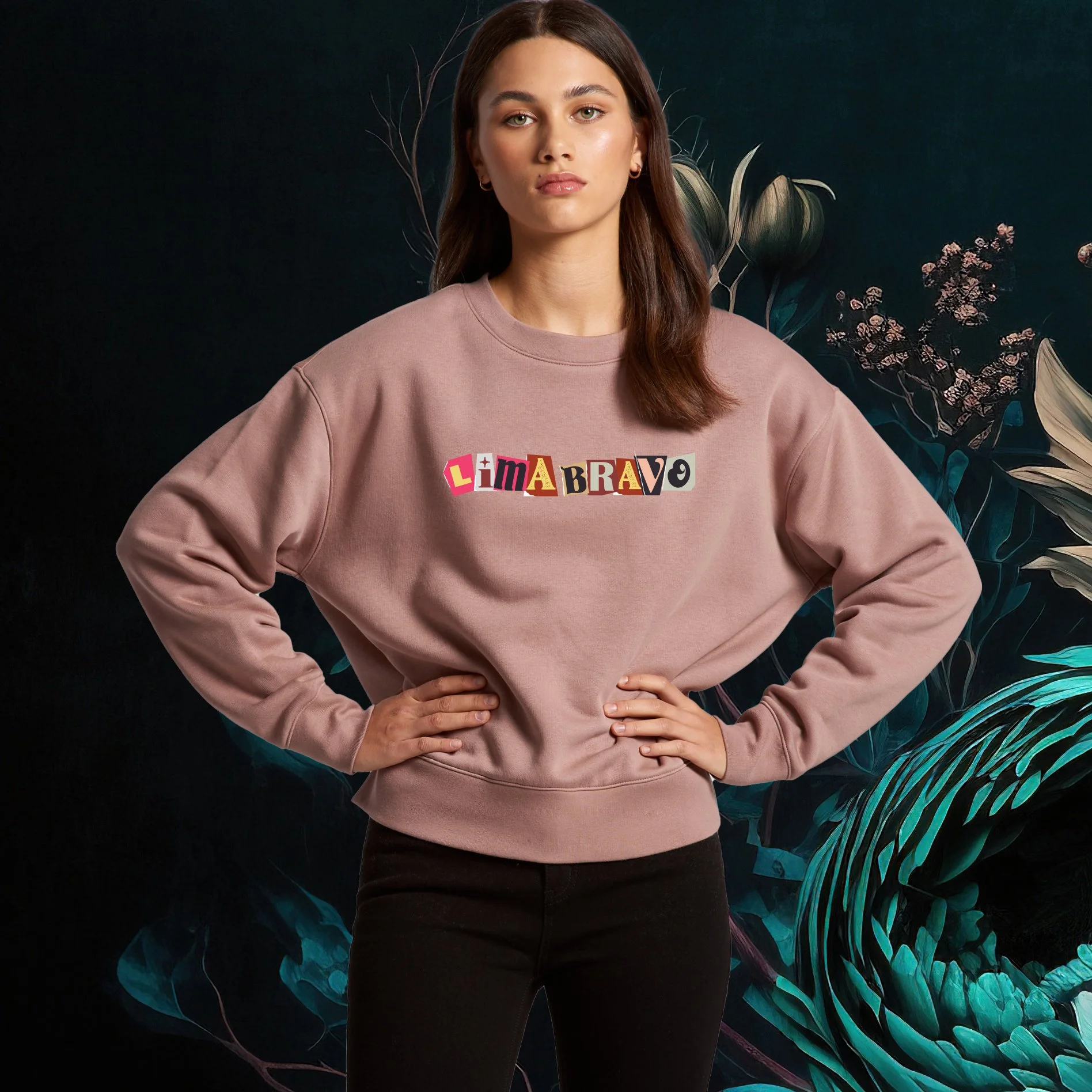 A young woman with brown hair and green eyes standing in front of a dark background with tropical leaves. She is wearing a pink sweatshirt with the words 'Lima Bravo' in a ransom note style font.