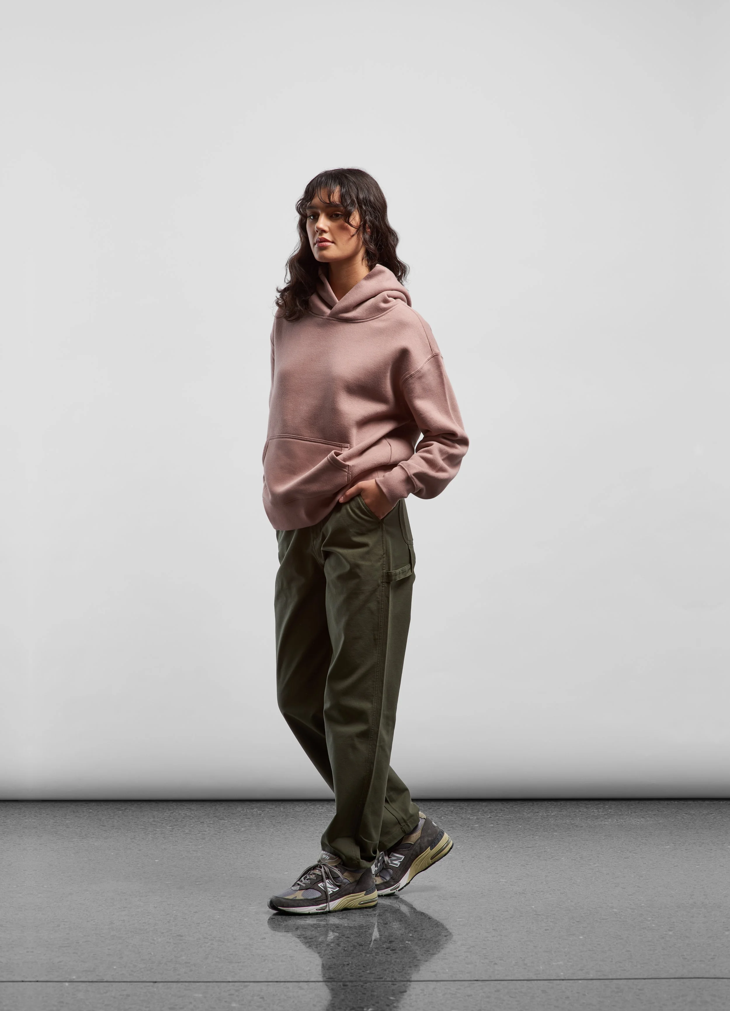 A woman in a pink hoodie and green pants standing with her hands in her pockets, wearing gray sneakers, against a plain white background.