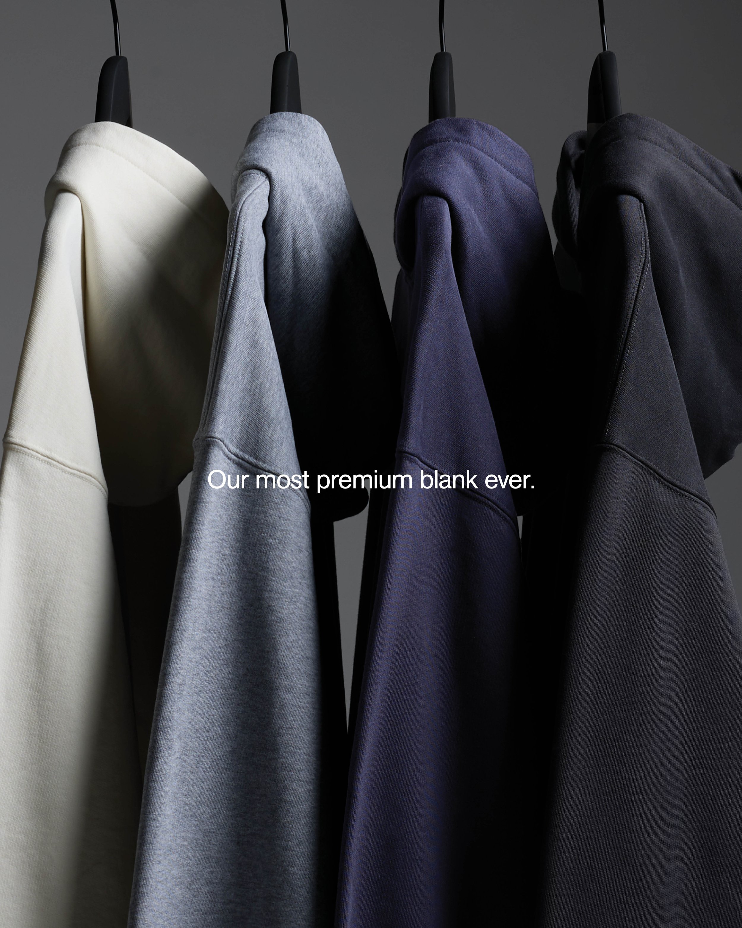 Four sweatshirts hanging on black hangers against a dark grey background, in cream, light grey, navy, and black colors.