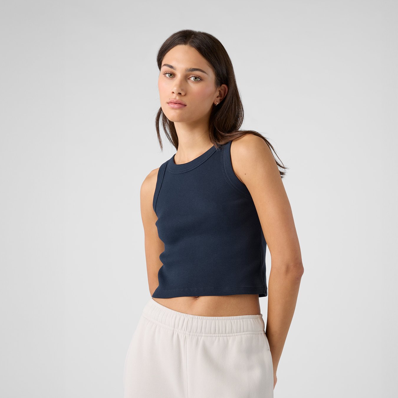 A young woman with medium-length brown hair wearing a sleeveless navy blue crop top and white high-waisted pants standing against a plain light gray background.