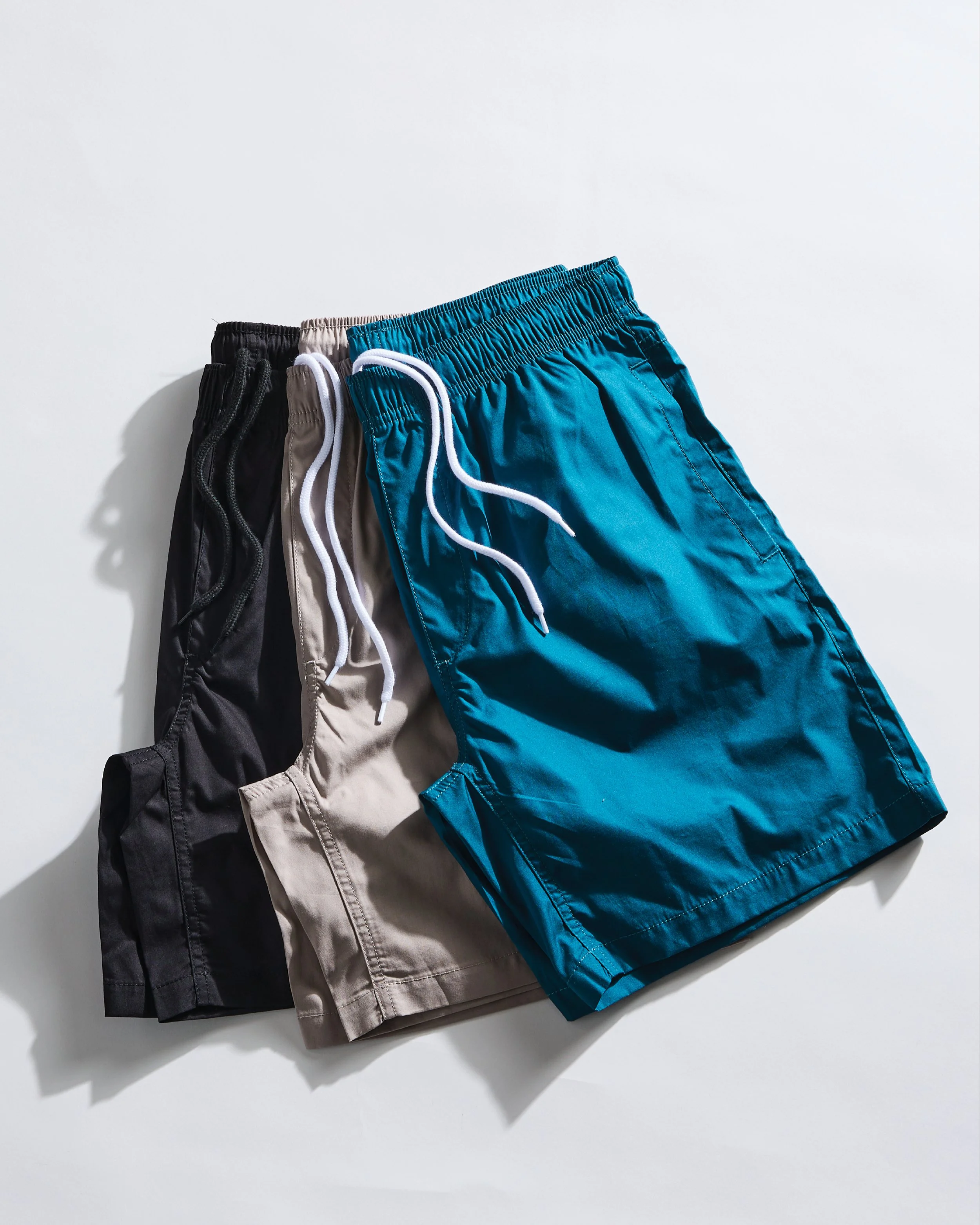 Three pairs of men's casual shorts in black, beige, and bright blue, folded and aligned side-by-side on a white surface.
