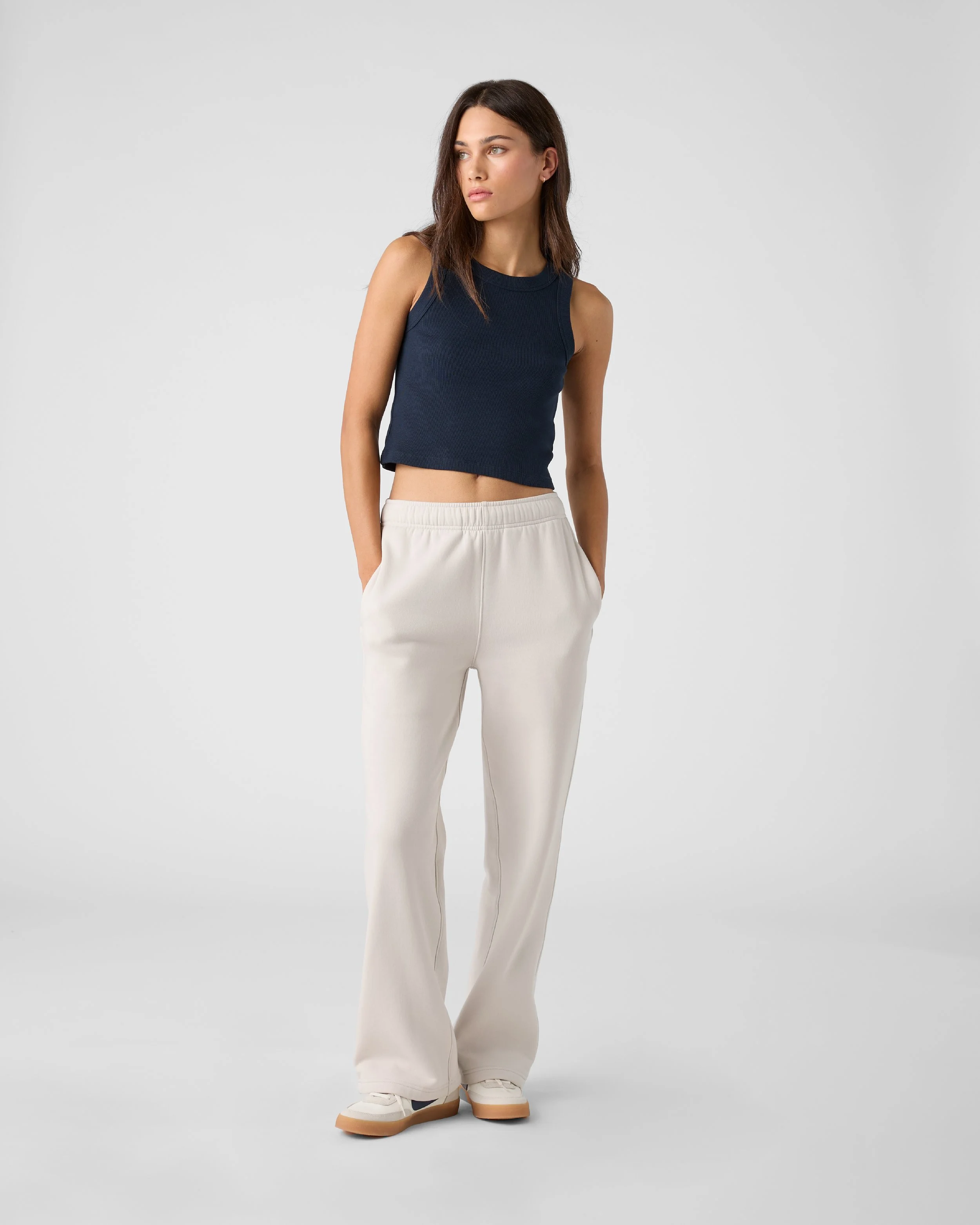 A young woman with shoulder-length brown hair in a casual outfit, standing against a plain light gray background. She is wearing a navy sleeveless crop top, light-colored wide-leg pants, and white sneakers with tan soles. She has a neutral expression