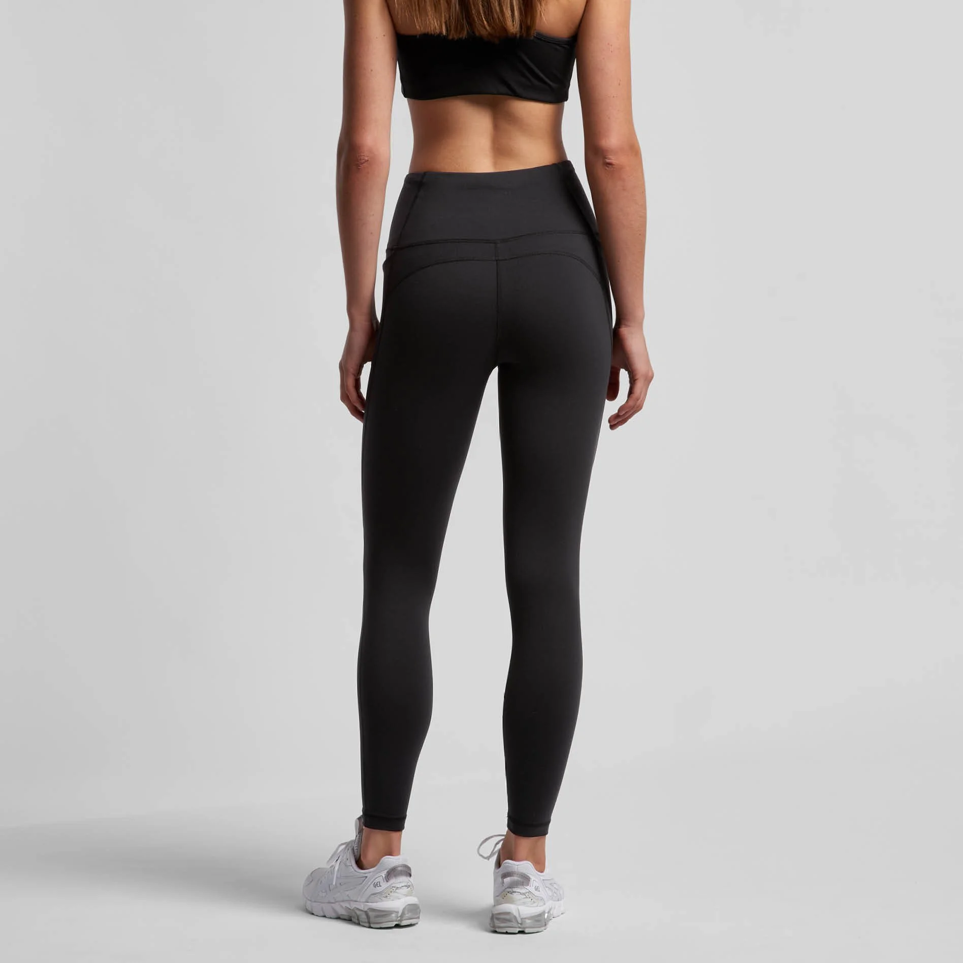 Back view of a woman wearing black athletic leggings, a black sports bra, and white sneakers, standing against a gray background.