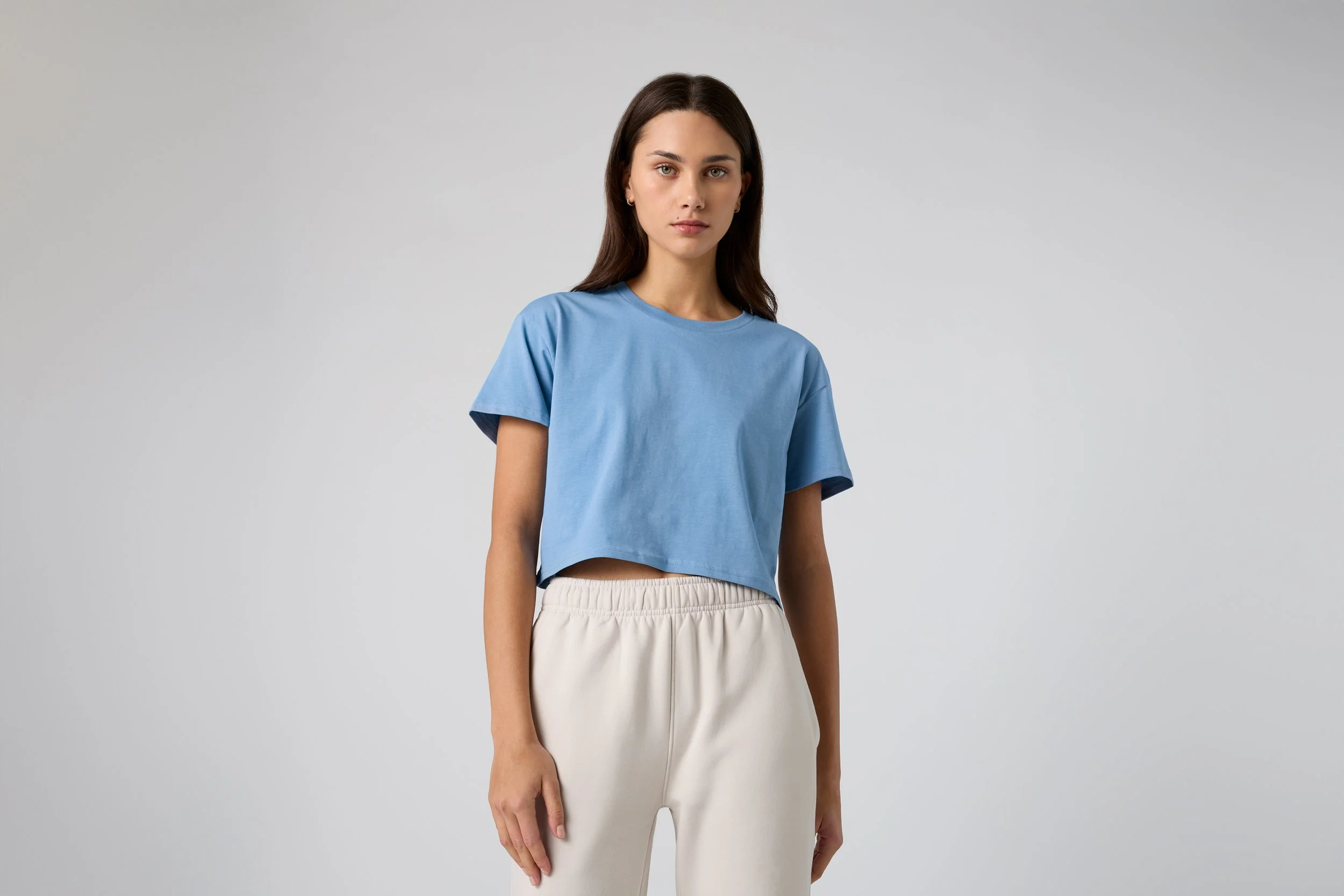 A young woman with long dark hair, wearing a light blue cropped T-shirt and beige pants, standing against a plain gray background.