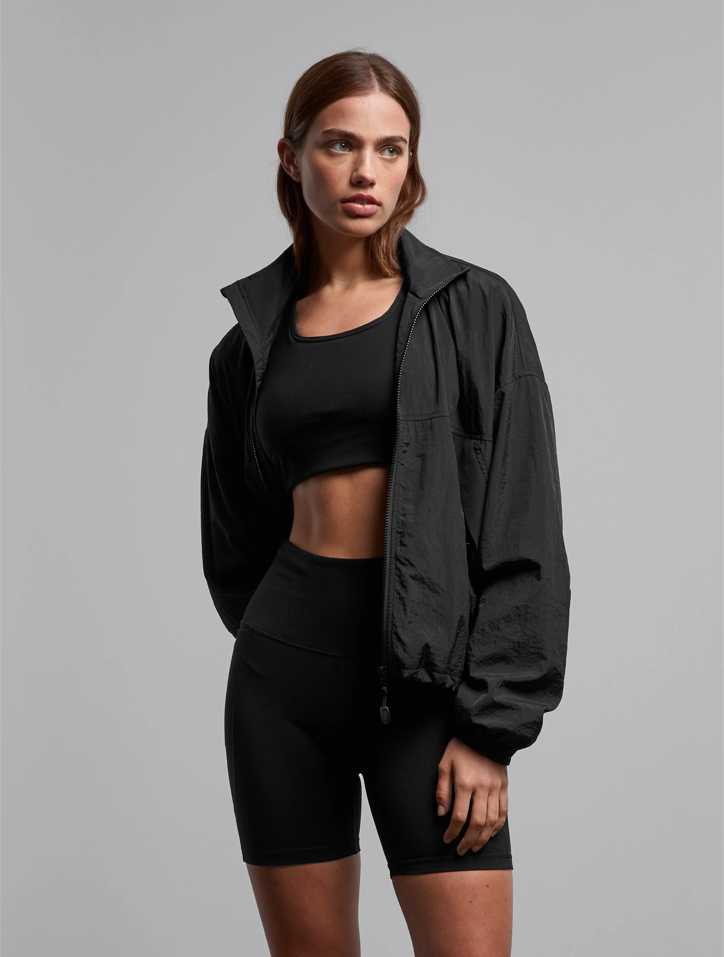 A woman with medium-length brown hair wearing a black zip-up jacket over a black crop top, and black shorts, standing against a plain gray background.