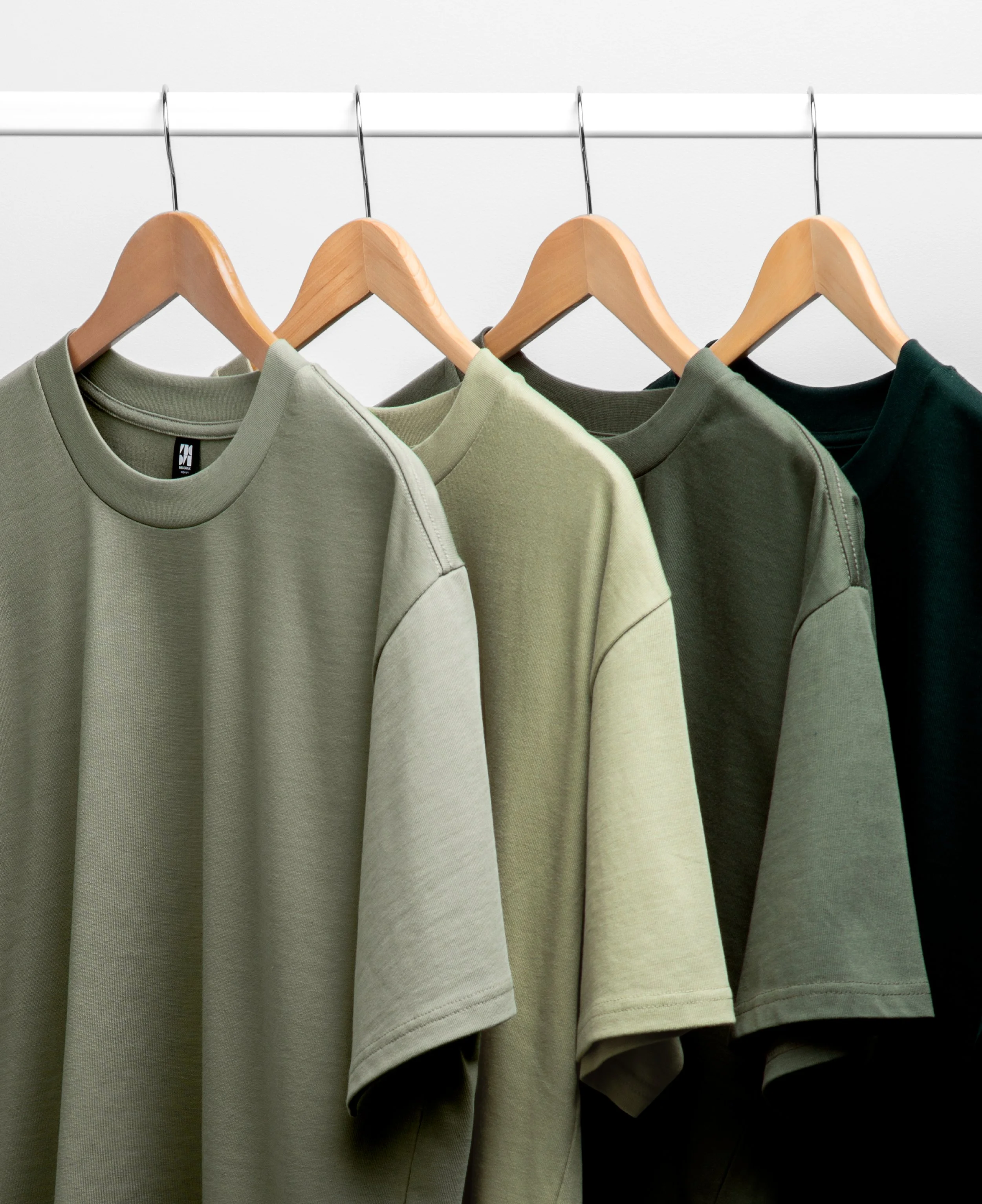Four plain crewneck sweatshirts hanging on wooden hangers on a metal clothing rack, varying in shades of green, from light to dark.
