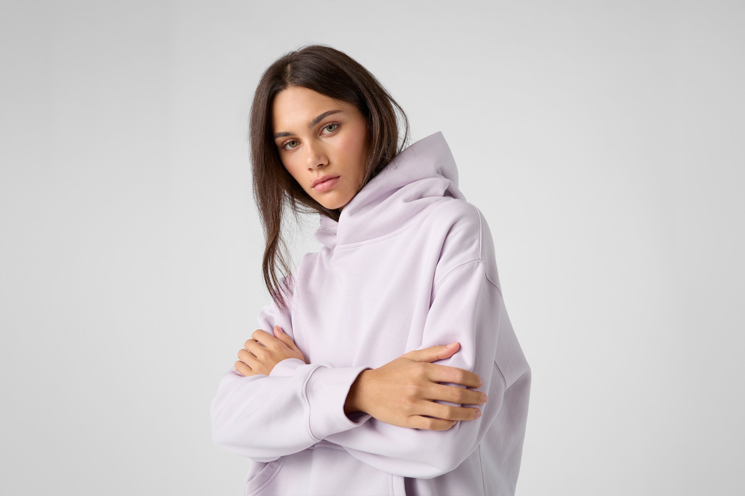 A young woman with brown hair and light eyes wearing a light pink hoodie, standing against a plain light gray background with her arms crossed, looking at the camera with a neutral expression.
