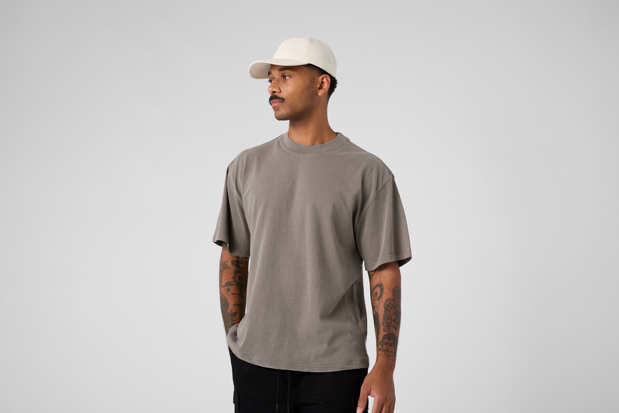 A man wearing a light gray oversized T-shirt, black pants, a white baseball cap, standing against a plain light gray background.