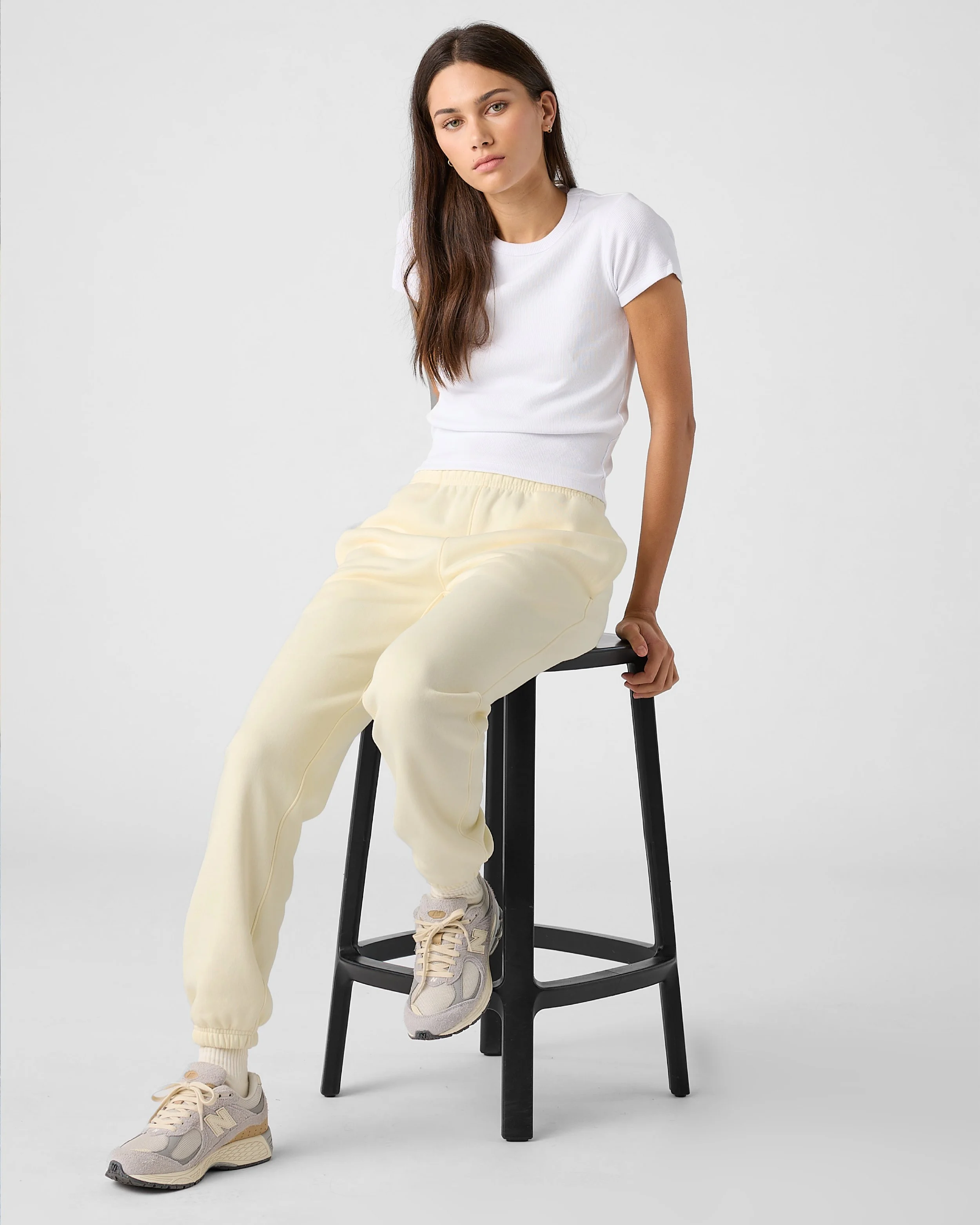 A young woman with long brown hair, wearing a white t-shirt, light yellow baggy sweatpants, and beige sneakers, sitting on a black stool against a plain white background.