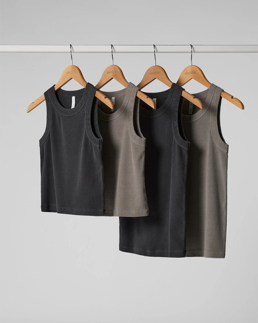 Four sleeveless tank tops hanging on wooden hangers against a plain light gray background, with two black and two gray.