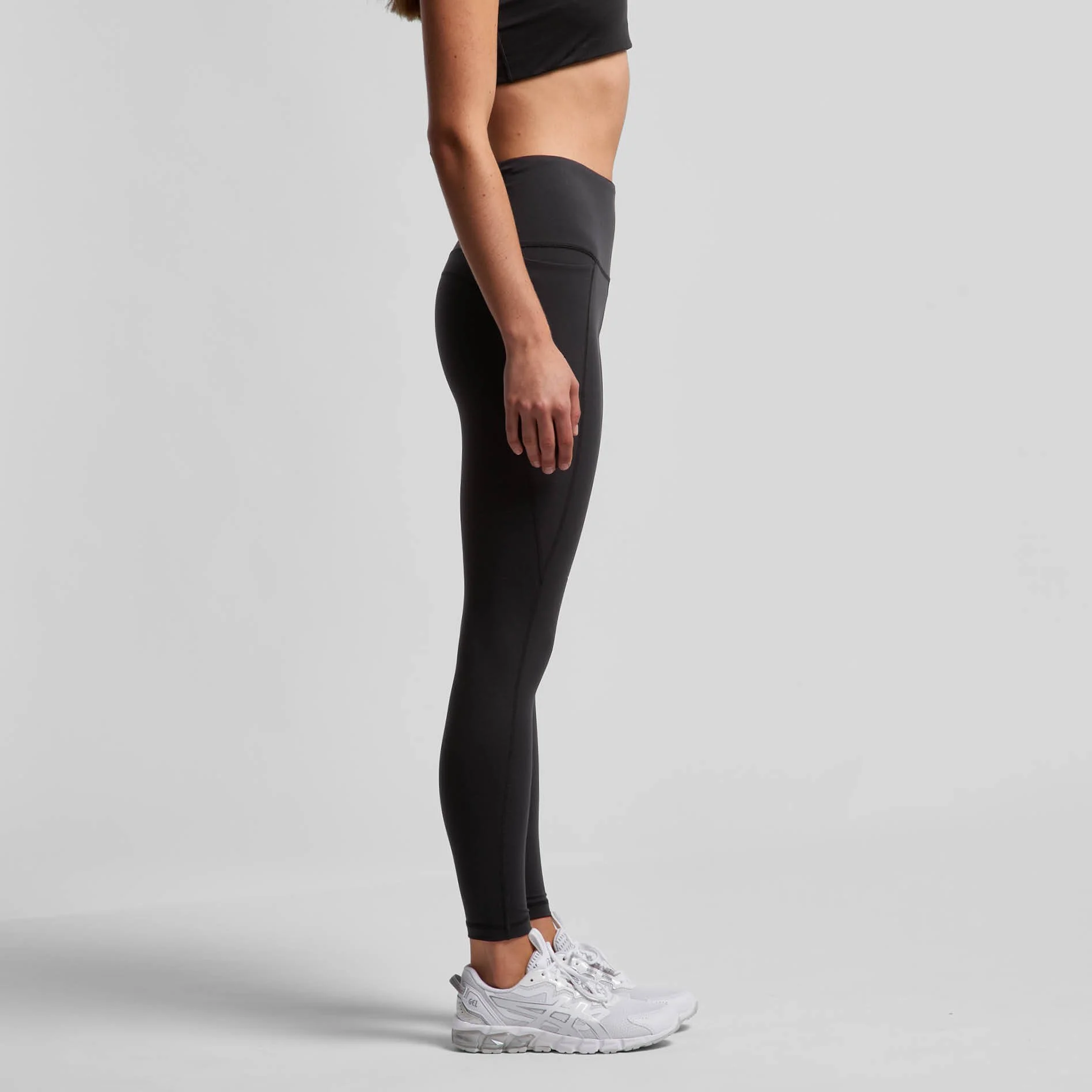 Side view of a woman wearing black athletic leggings, a black top, and white sneakers, standing against a light gray background.
