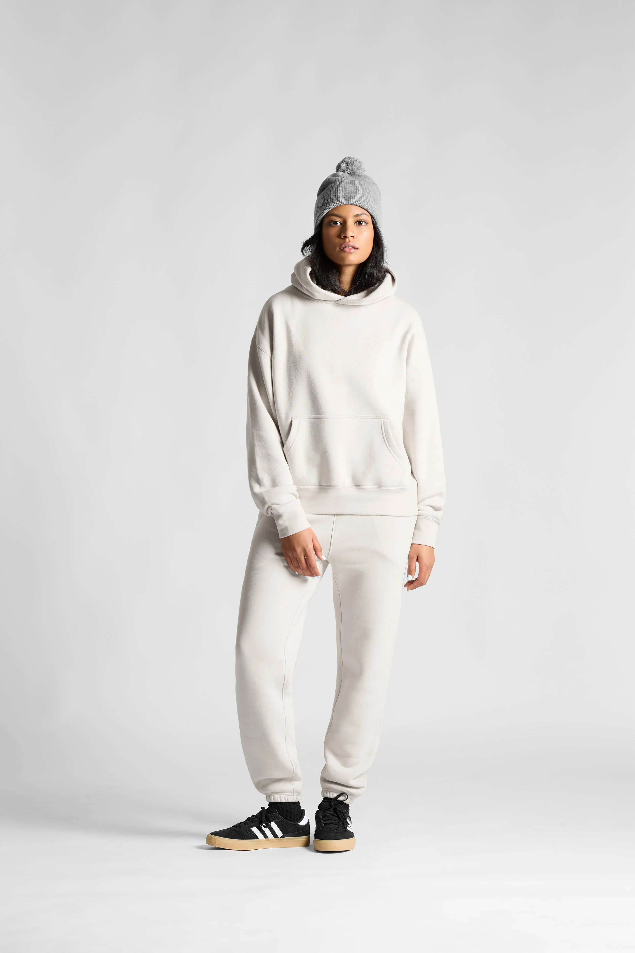 A young woman standing against a plain light gray background, wearing a cream-colored hoodie, matching sweatpants, a gray knit beanie with a pom-pom, and black sneakers with white stripes.