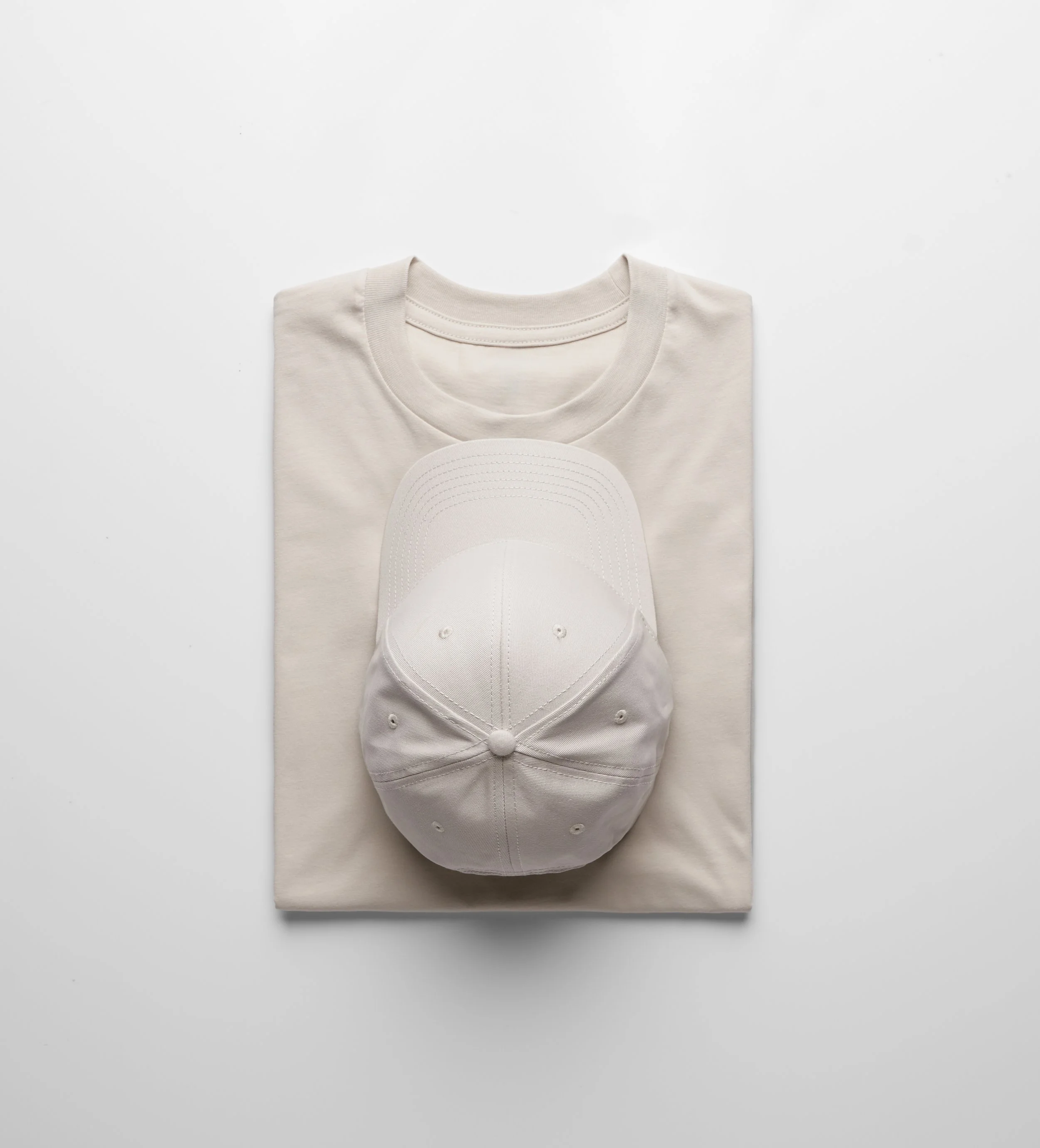 A folded beige t-shirt with a baseball cap on top, both on white packaging paper.