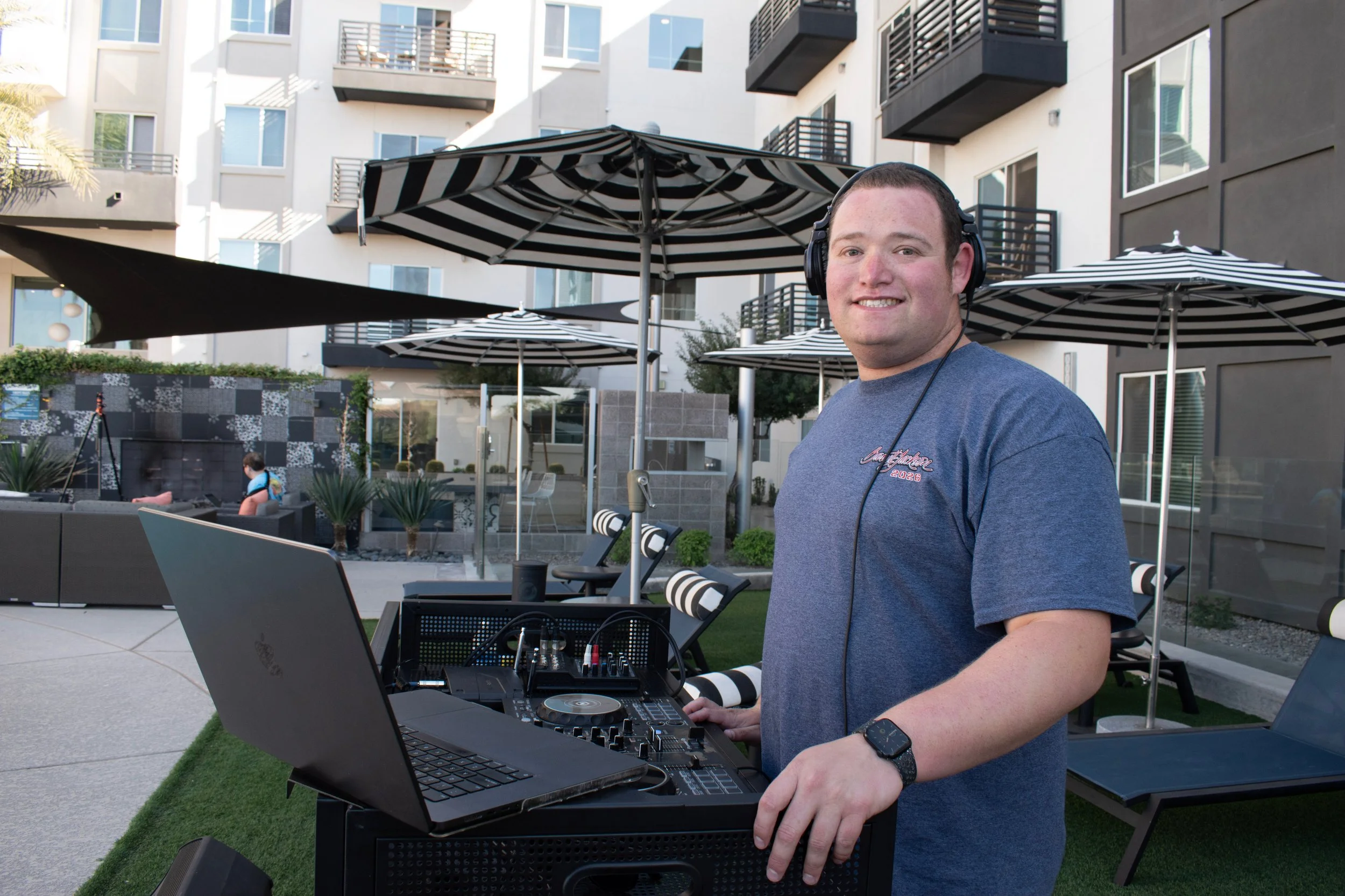A smiling man wearing headphones and a blue t-shirt standing next to DJ equipment and a laptop outdoors on a patio with black and white striped umbrellas.