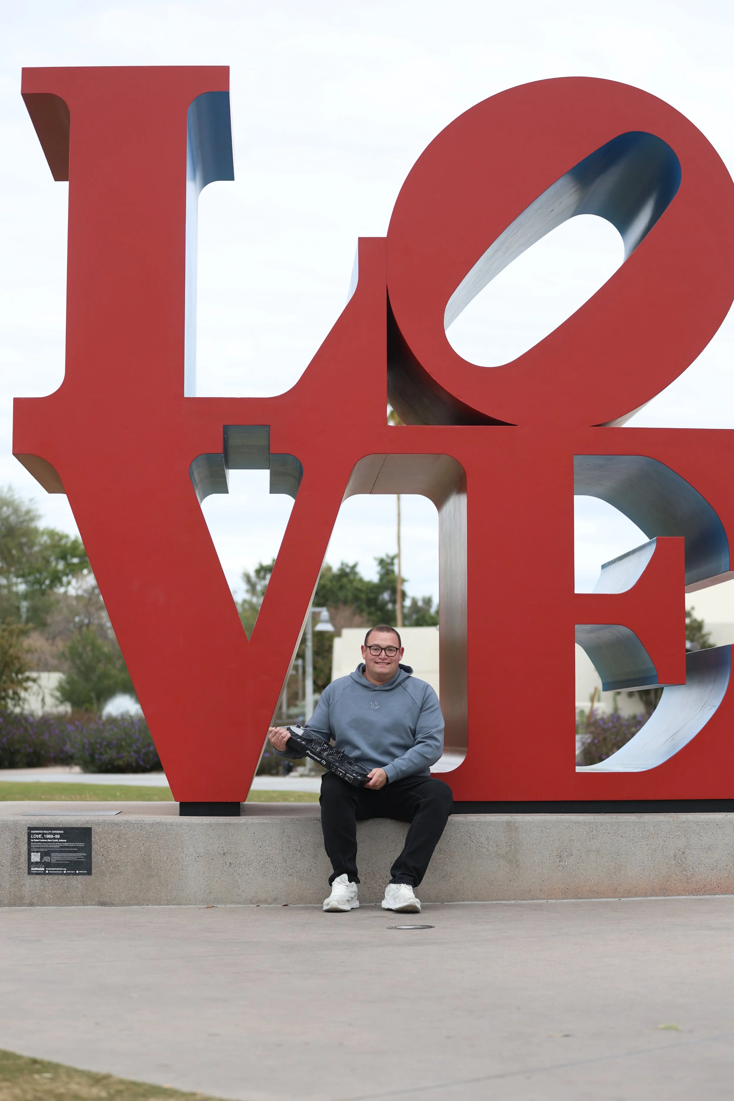 A young man sitting in front of a large red sculpture spelling the word LOVE, holding a person's hands. The sculpture is outdoors with trees and buildings in the background.