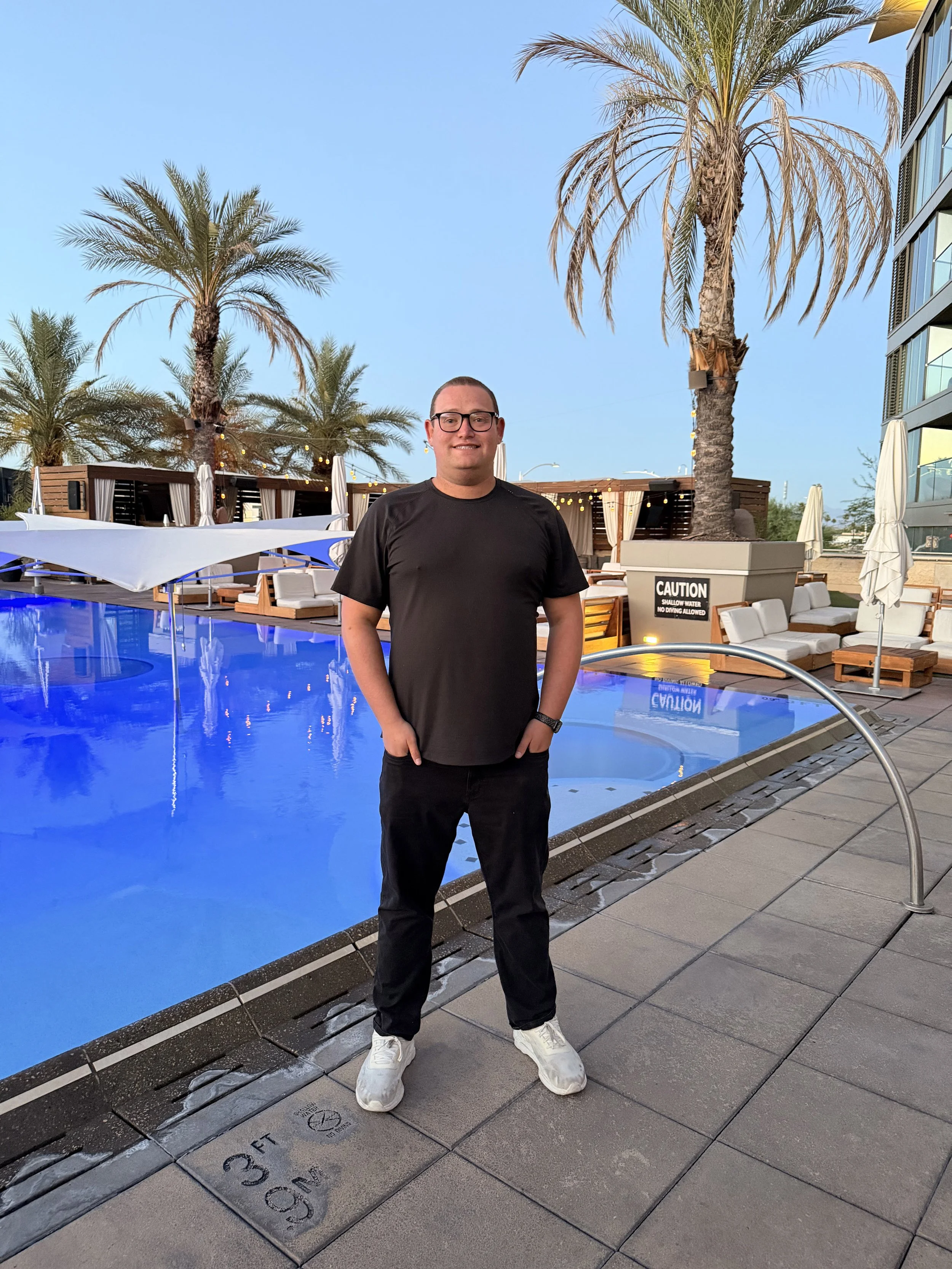 A man standing near a pool with palm trees and cabanas in the background, wearing a black t-shirt, black pants, white sneakers, and glasses, during the daytime.