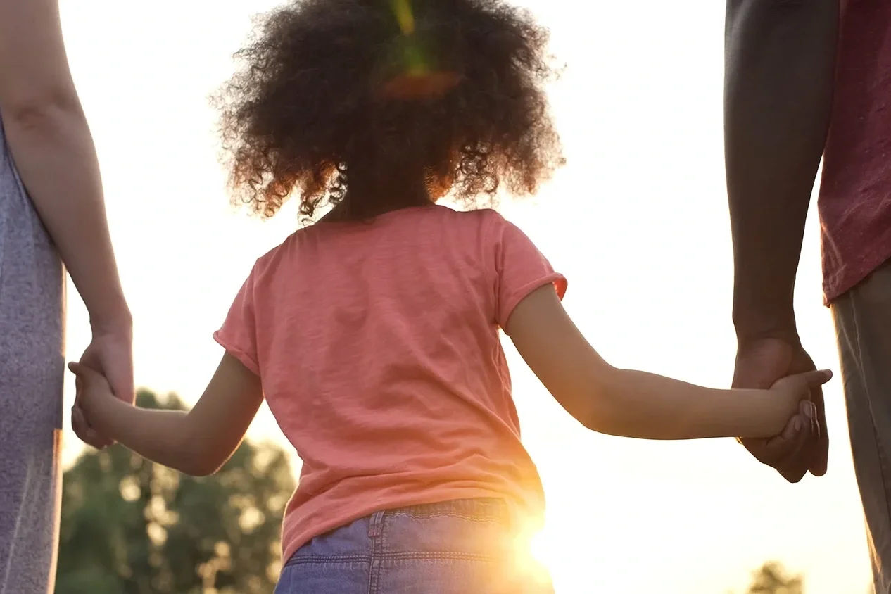 Child holding hands with two adults while walking outdoors at sunset.