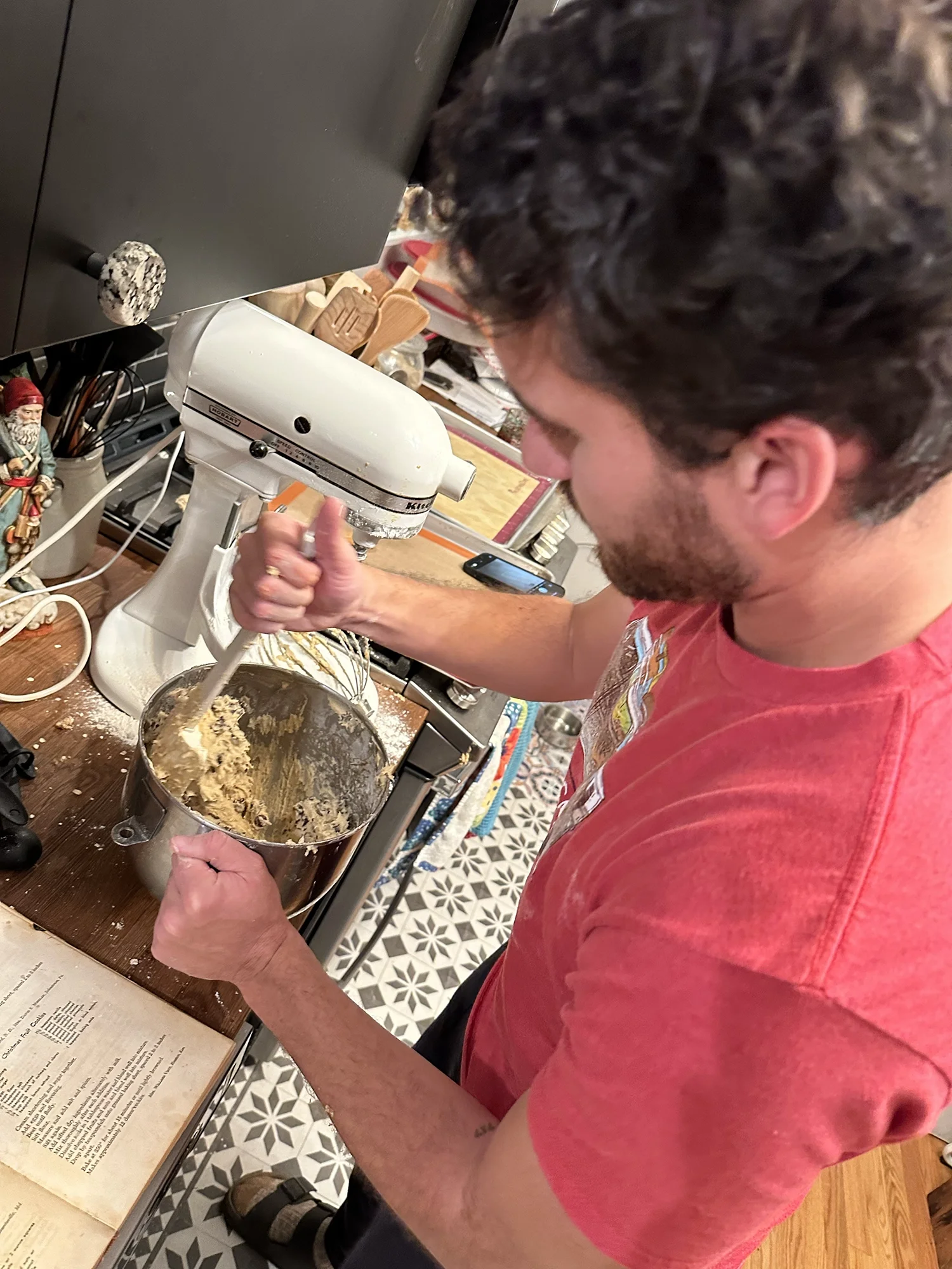 Man mixing cookie dough in a stand mixer in a home kitchen, with a recipe book open nearby.