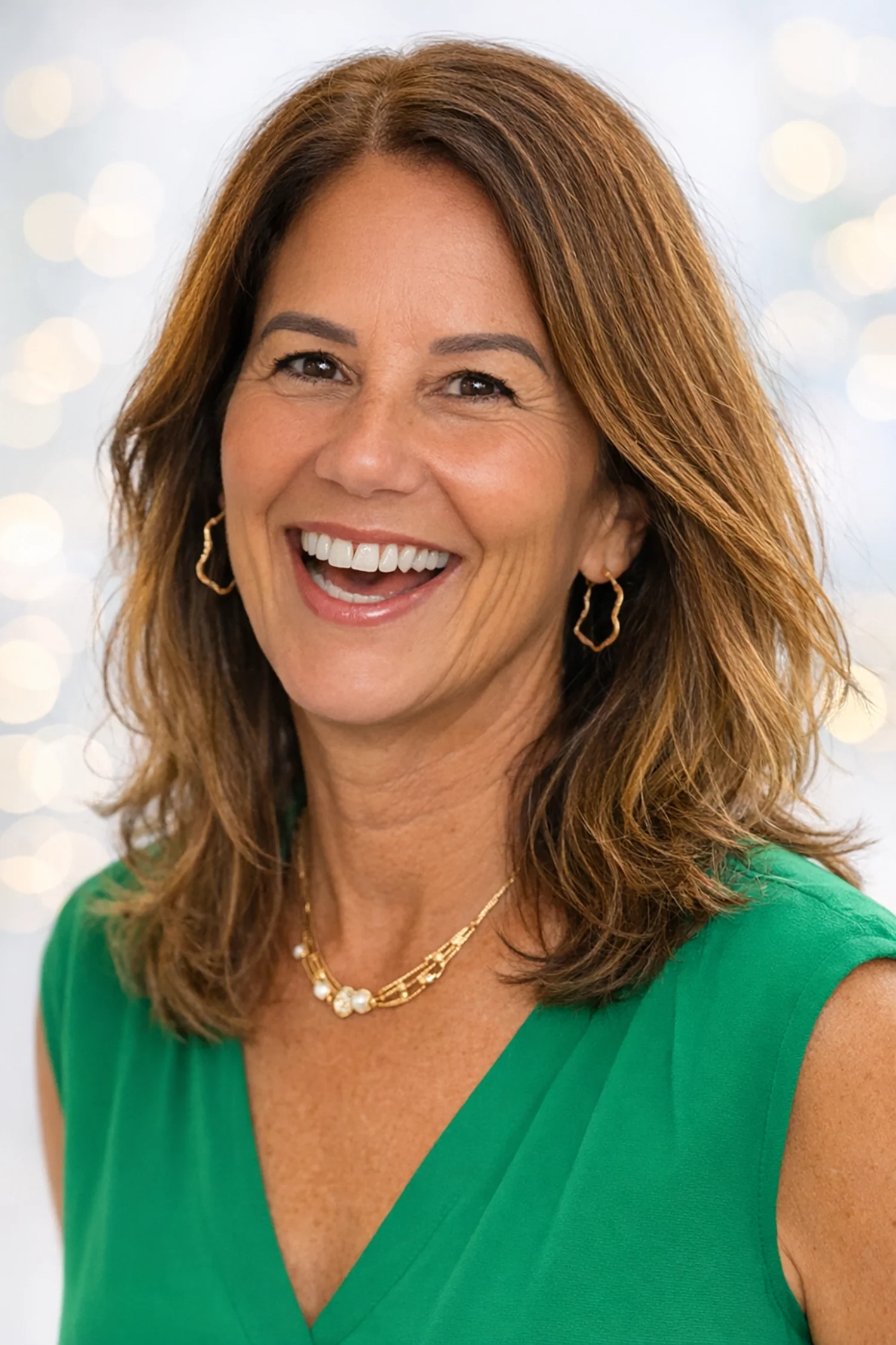 Smiling woman wearing a green top and gold jewelry, posing for a professional headshot against a softly blurred background.