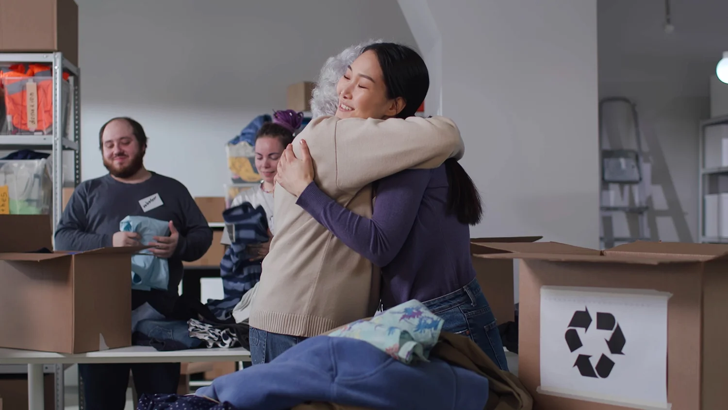 Two women hugging in a donation center while volunteers sort clothing and pack boxes in the background.