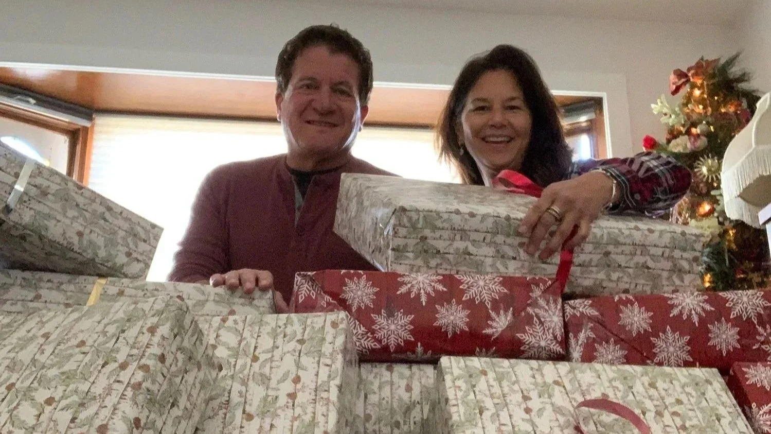 Two people smiling while holding stacked wrapped holiday gifts near a decorated Christmas tree.