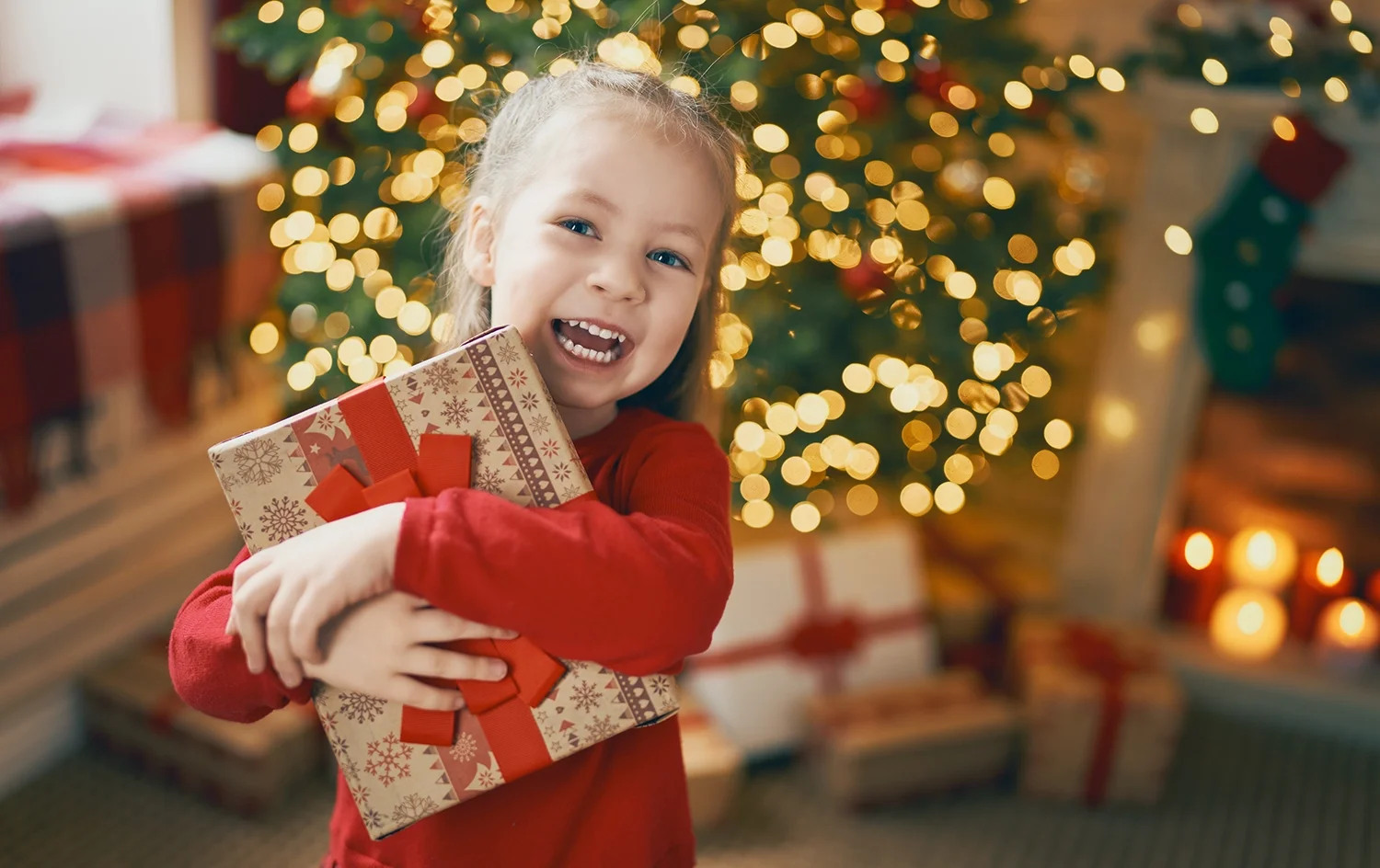 Smiling young girl holding a wrapped Christmas gift in front of a decorated tree with lights and presents.