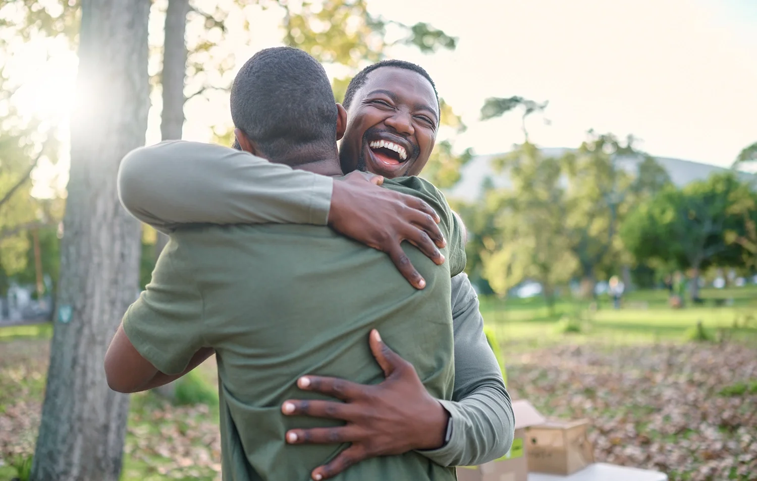 Two men smiling and hugging outdoors in a park, showing friendship and support.