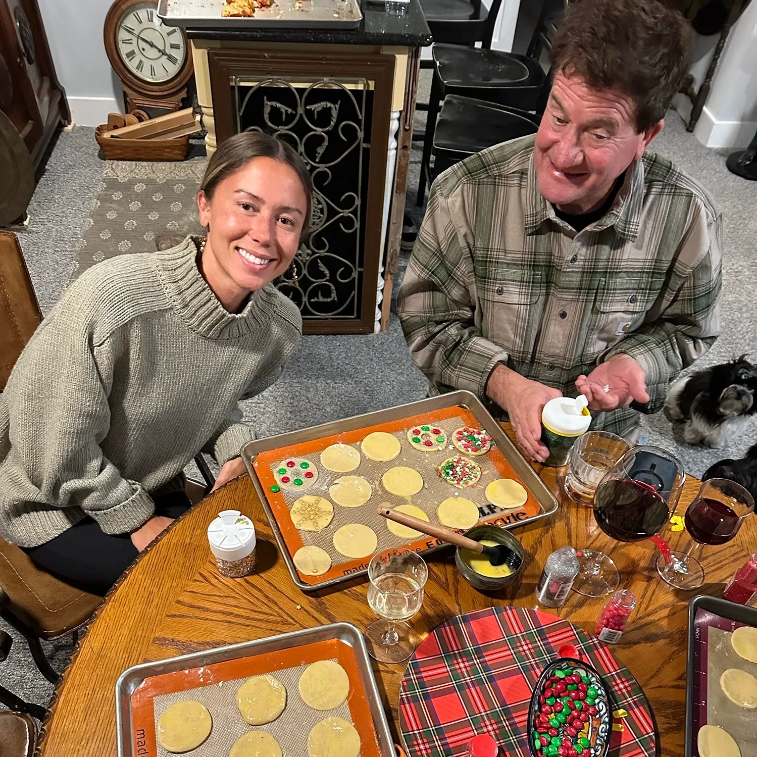 Two people decorating holiday cookies together at a kitchen table filled with sprinkles, dough, and baking trays.