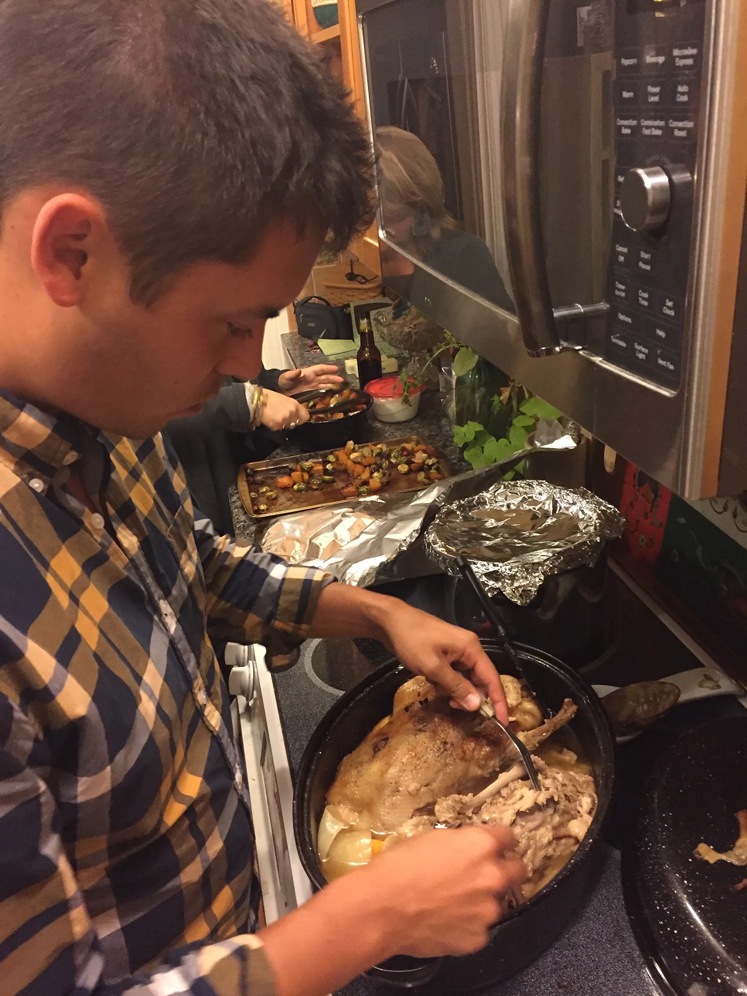 Man carving cooked chicken in a kitchen while others prepare roasted vegetables in the background.