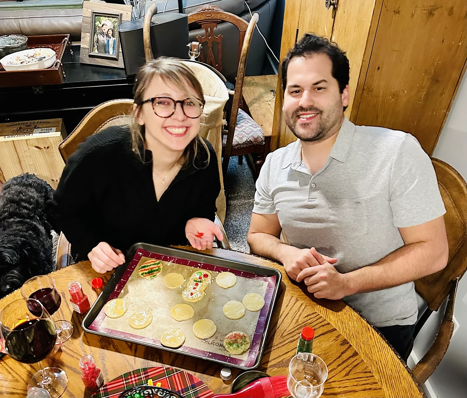 Two people smiling at a table while decorating holiday cookies with sprinkles and icing.