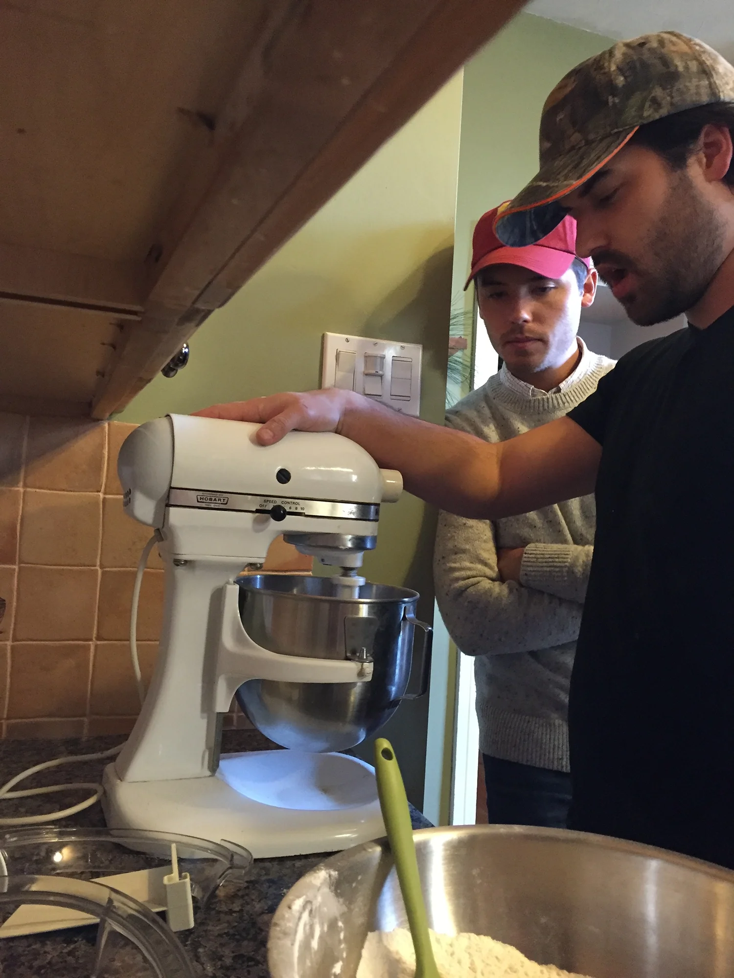 Two men using a stand mixer in a kitchen, one operating the machine while the other watches.