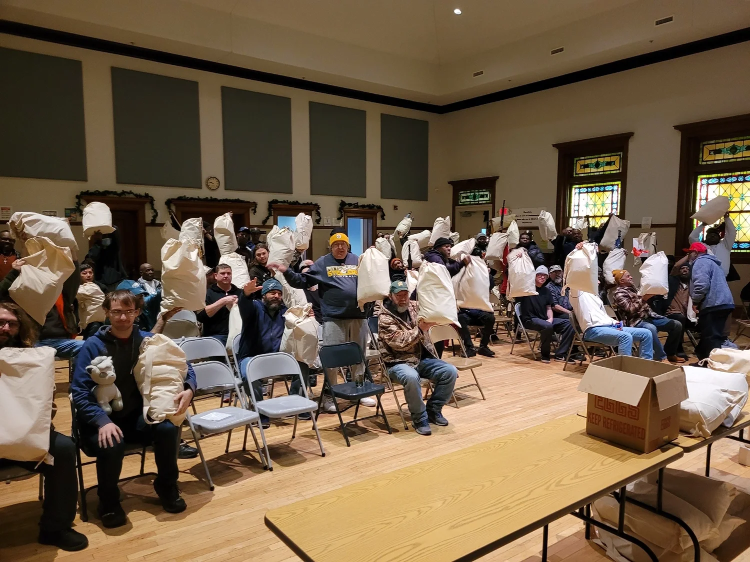 Group of people seated in a community hall holding up donated bags during a distribution event.