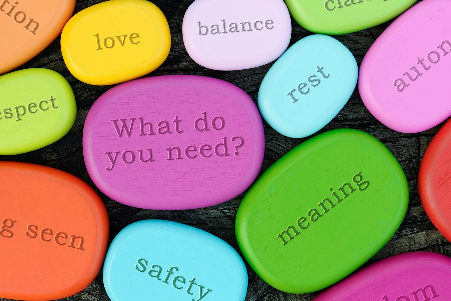 Colorful smooth pebbles with words like 'love,' 'balance,' 'respect,' 'safety,' 'rest,' 'trust,' 'autonomy,' 'meaning,' 'seen,' and 'what do you need?' engraved on them are scattered on a dark wooden surface.