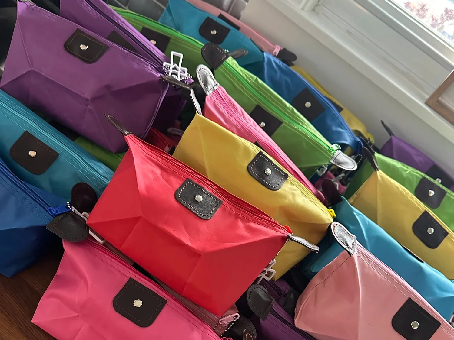 Colorful zippered pouches in purple, green, yellow, red, pink, blue, and black with brown and silver accents, arranged on a table near a window