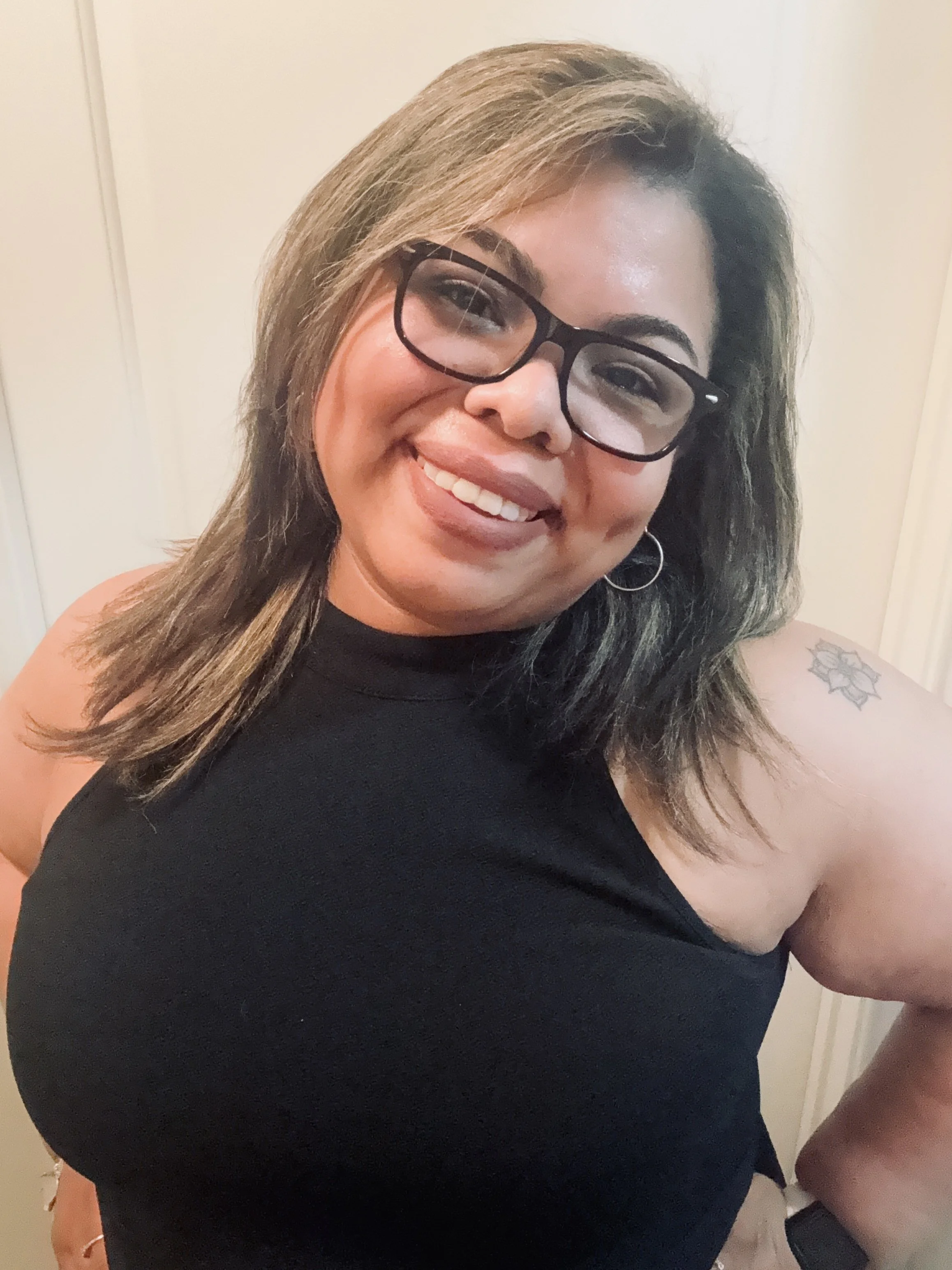A woman smiling, wearing black glasses and a black top, with shoulder-length brown hair. She has a small tattoo on her arm and hoop earrings.