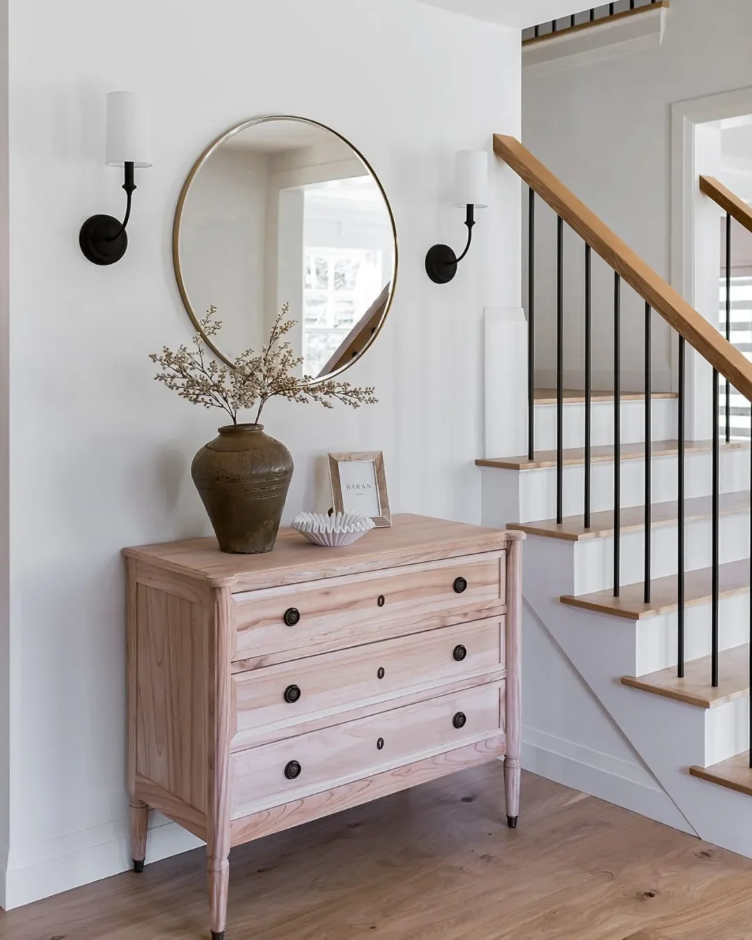 A wooden dresser with four drawers, decorated with a large vase with dried branches, a framed picture holder, and a small white bowl, positioned under a round mirror flanked by two black wall-mounted sconces with white shades, near a staircase with wooden steps and black metal railings.