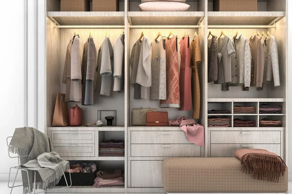 An organized wardrobe with hanging clothes and folded items, with a chair draped with a blanket and a cushioned bench in front.