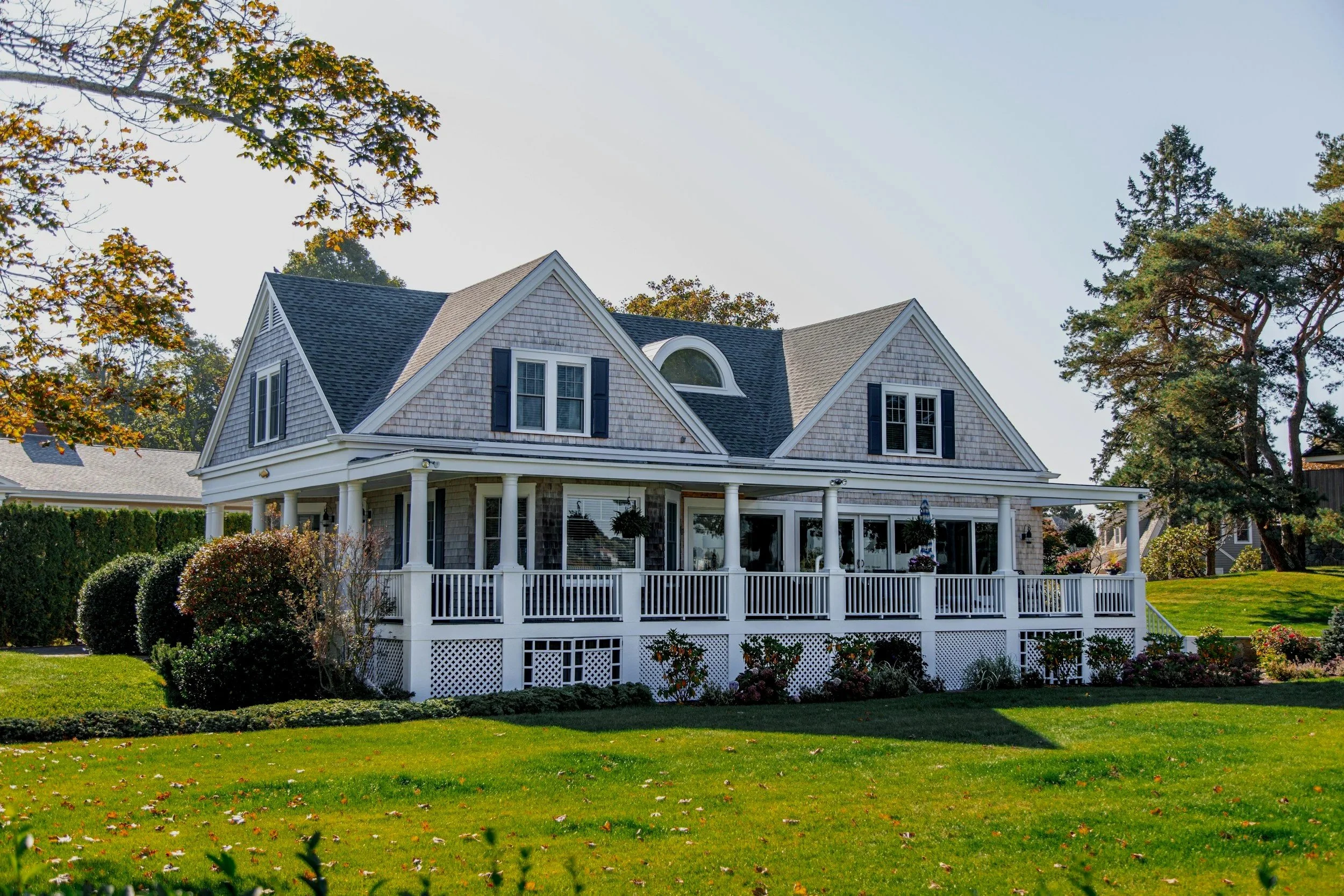 A large two-story house with a porch, multiple windows, and a hip roof, surrounded by a well-maintained yard with grass, bushes, and trees.