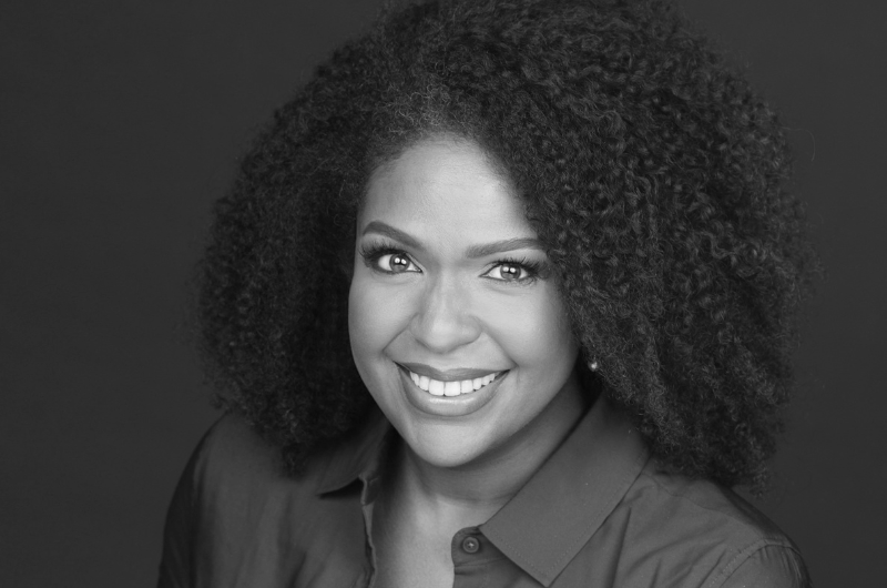 A black and white headshot of a woman with voluminous curly hair smiling warmly, wearing a collared shirt.
