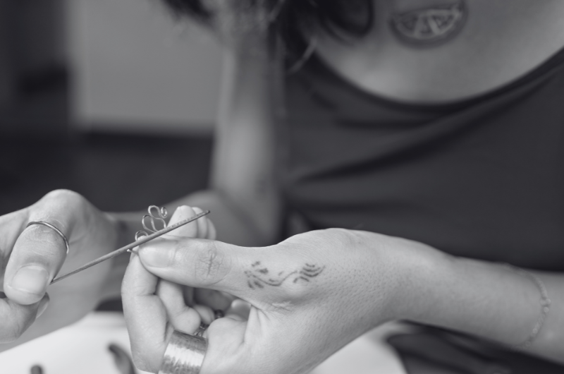 Close-up of hands holding a small metal tool and threading a needle into a thin wire, with a tattooed wrist and a blurred person in the background.