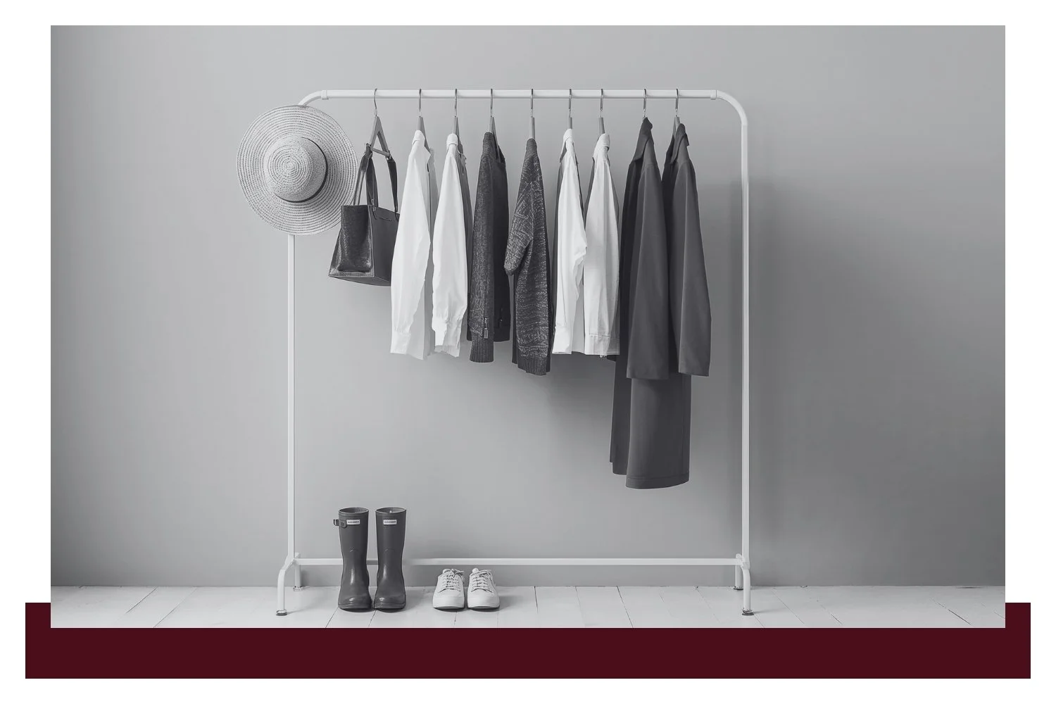 A minimalist clothing rack with neatly hung coats and shirts in dark and light colors, plus boots and small items on the floor against a plain gray wall.