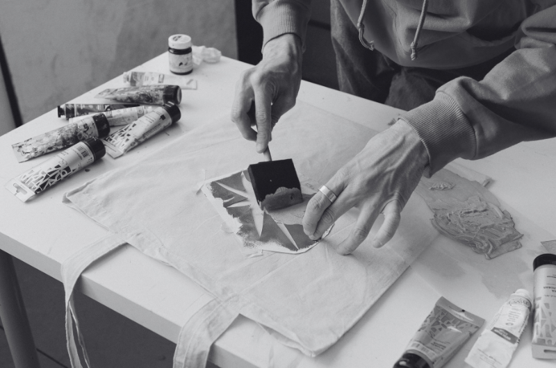 A person is using a sponge to apply paint to a fabric bag, with tubes of paint scattered on the table.
