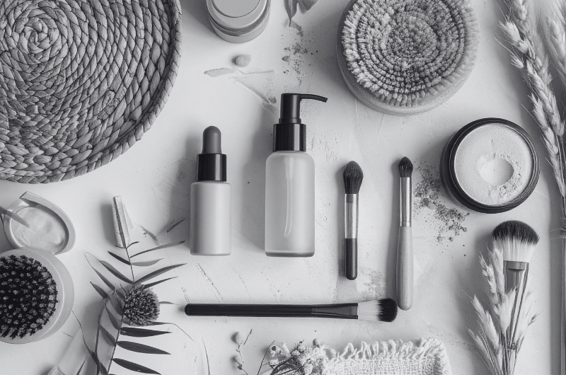 A flat lay of various makeup products including foundation bottles, brushes, powder, and a woven accessory.