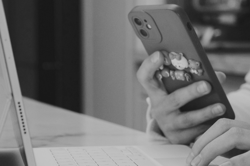 A person holding a smartphone with a patterned case while using it at a desk, with a laptop keyboard visible in the foreground and a blurred background.
