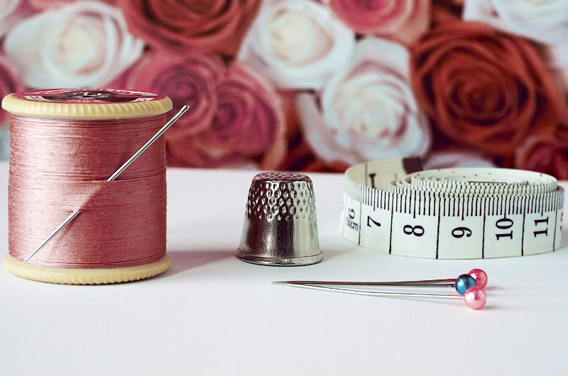 A spool of pink thread with a needle, a silver thimble, and a measuring tape are arranged on a white surface, with a blurred background of pink and white roses.