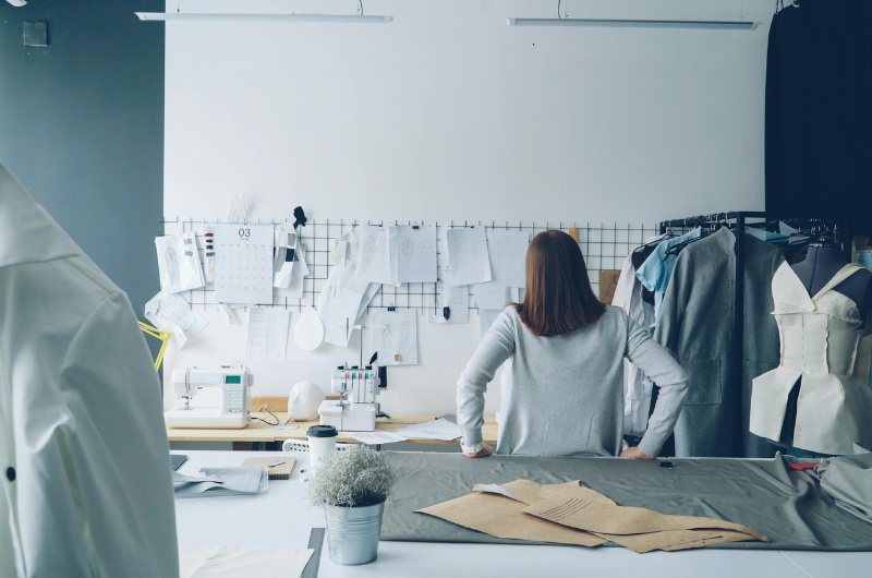 A person stands at a sewing table with fabric and patterns spread out, facing a wall covered in sketches and a calendar. A sewing machine and a dress form are visible on the table.