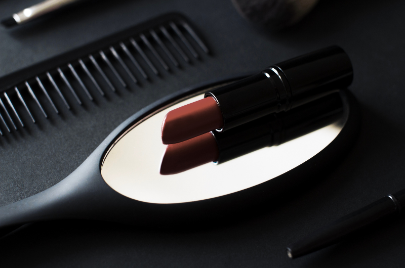 A dark background with a black comb, a makeup brush, a mirror reflecting a reddish-brown lipstick, and a black eyeliner pencil.