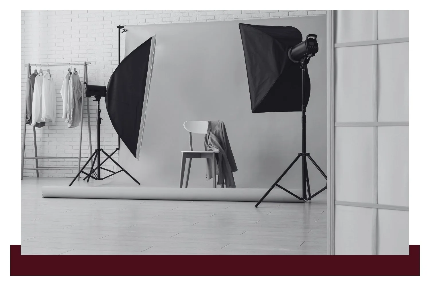 Black-and-white studio scene with two large softbox lights on tripods, a white backdrop, a chair and folded fabric on a set, and several hanging garments in the background.